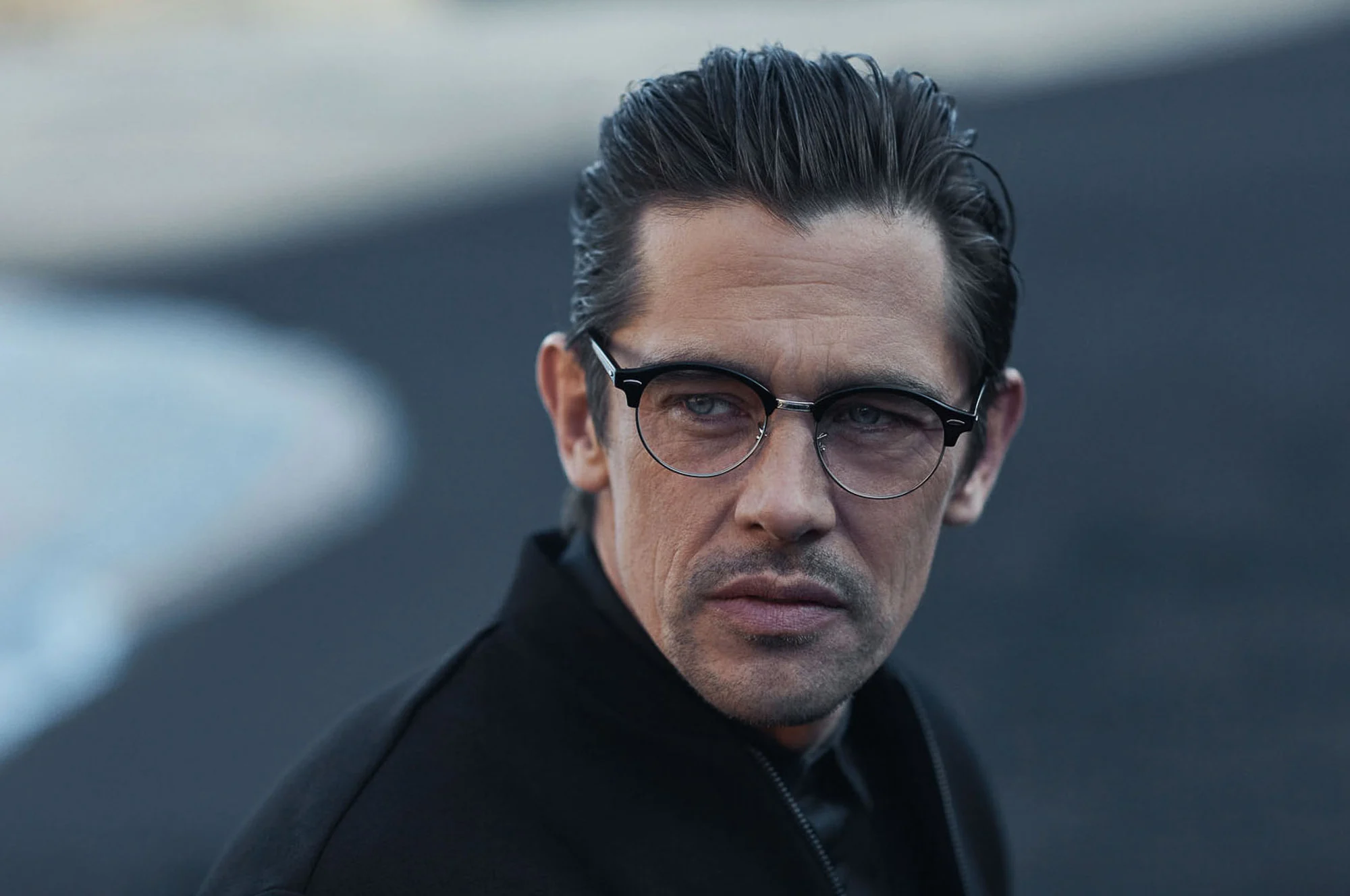 Man with slicked-back hair in browline glasses and black jacket on a dark beach, for Hiltl.
