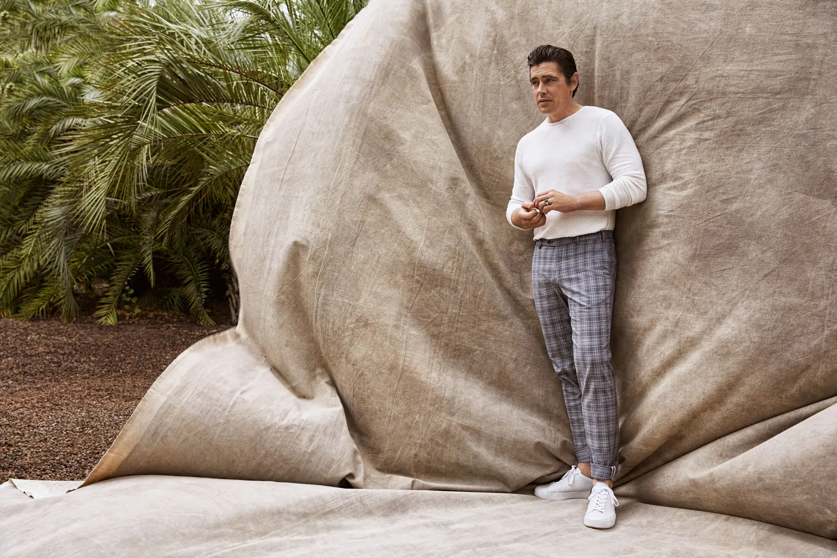 Man in white sweater, grey plaid trousers and white sneakers against a draped canvas backdrop beside palms, for Hiltl.