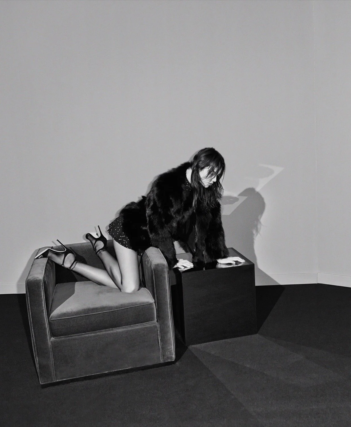 Black-and-white editorial of a model in a fur coat and strappy heels kneeling on a velvet armchair beside a black cube.