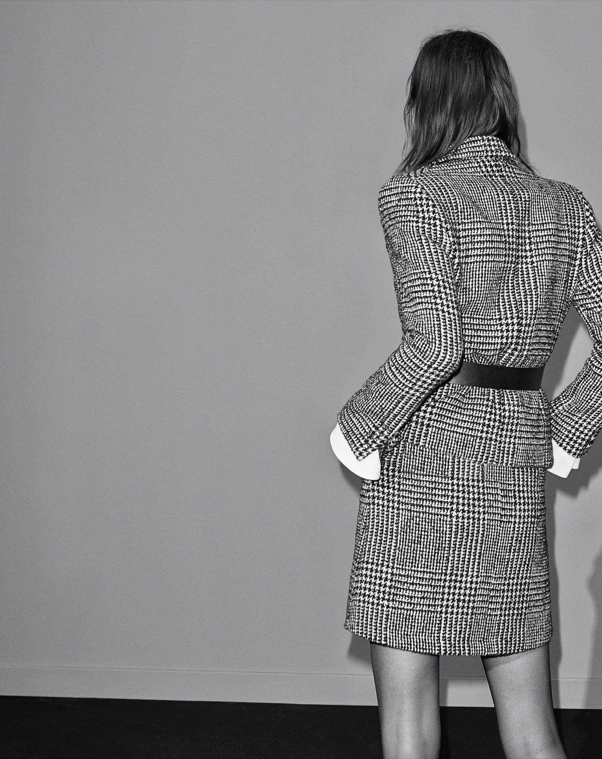 Black and white editorial of a model seen from behind in a belted houndstooth jacket and skirt against a grey wall.
