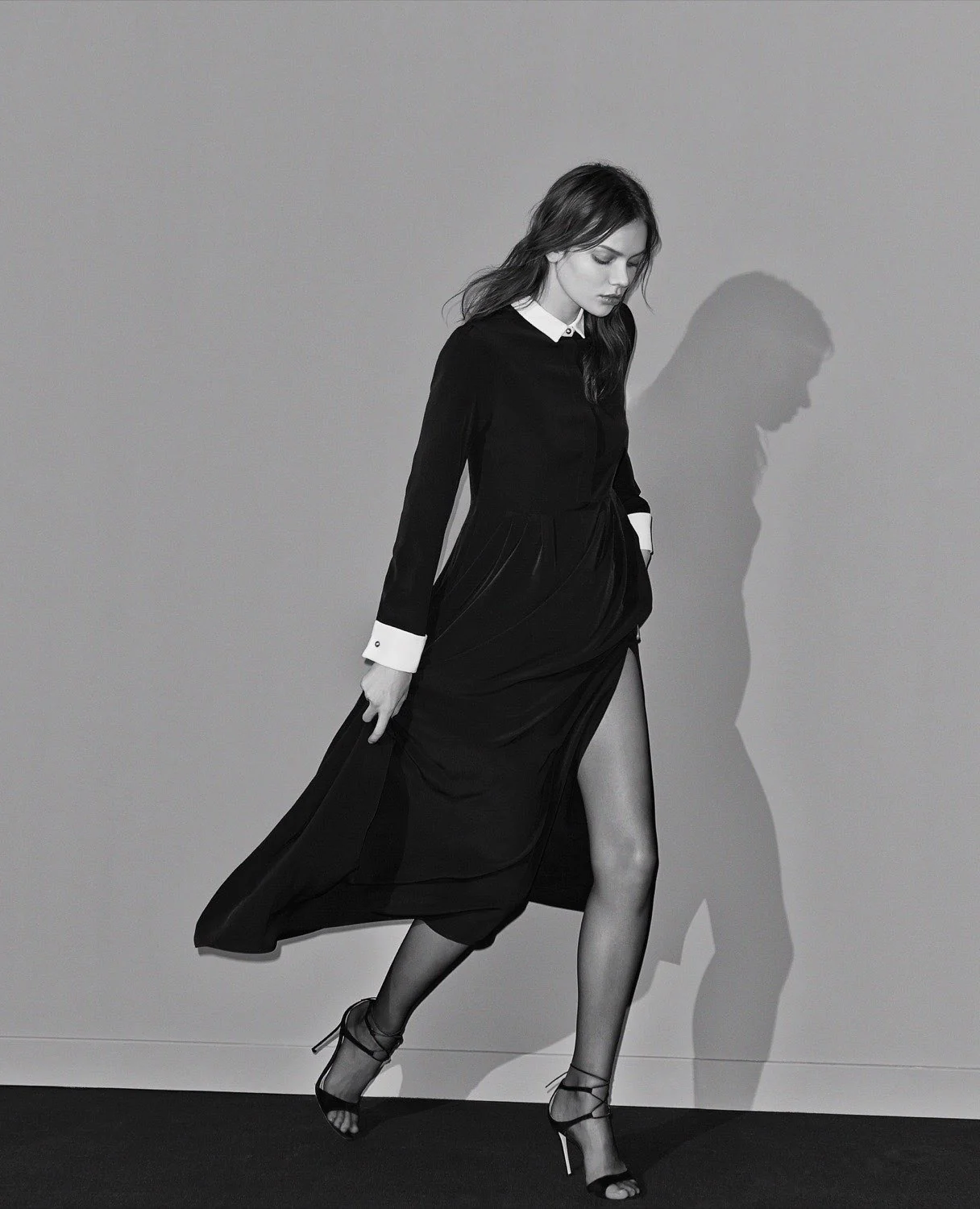 Black-and-white editorial of a model walking in a black collared shirt dress with high slit and strappy heels, for Academia.