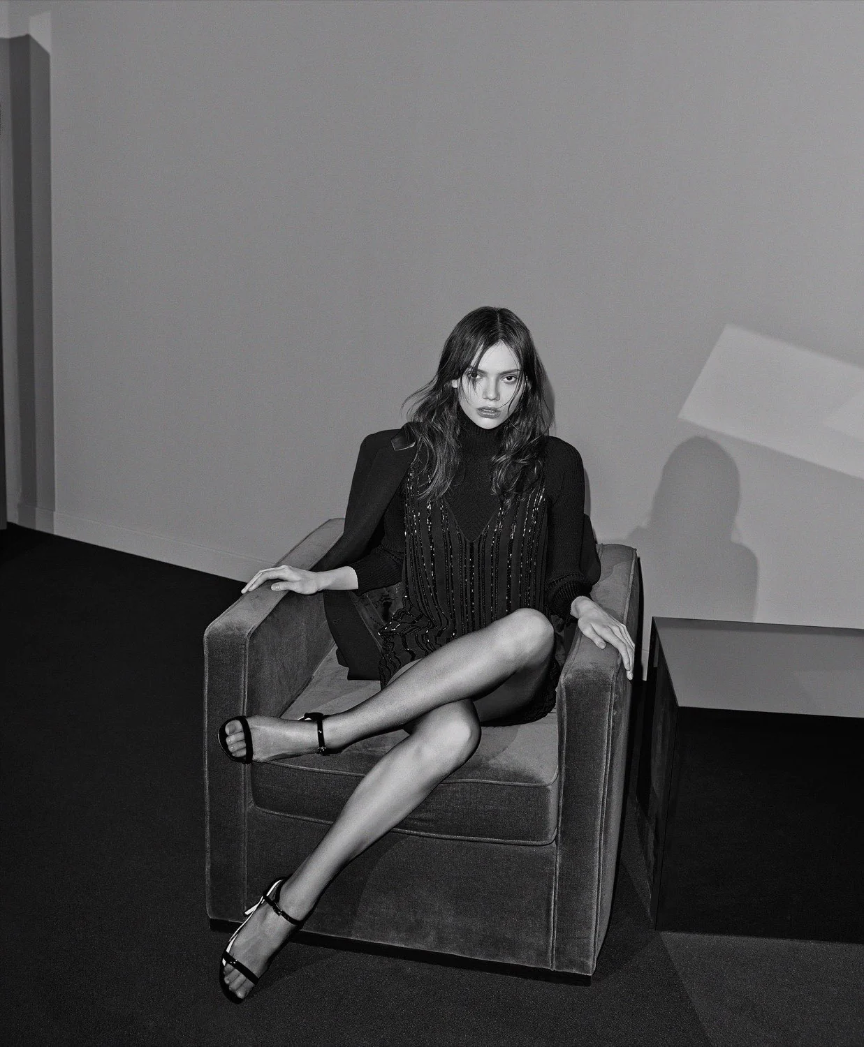 Black-and-white editorial of a model in a beaded black dress and strappy sandals, legs crossed on a velvet armchair.