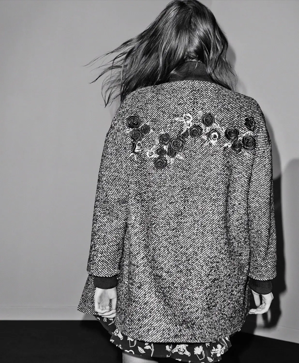Black and white shot of a model from behind in an oversized tweed coat with rose appliqués across the shoulders.