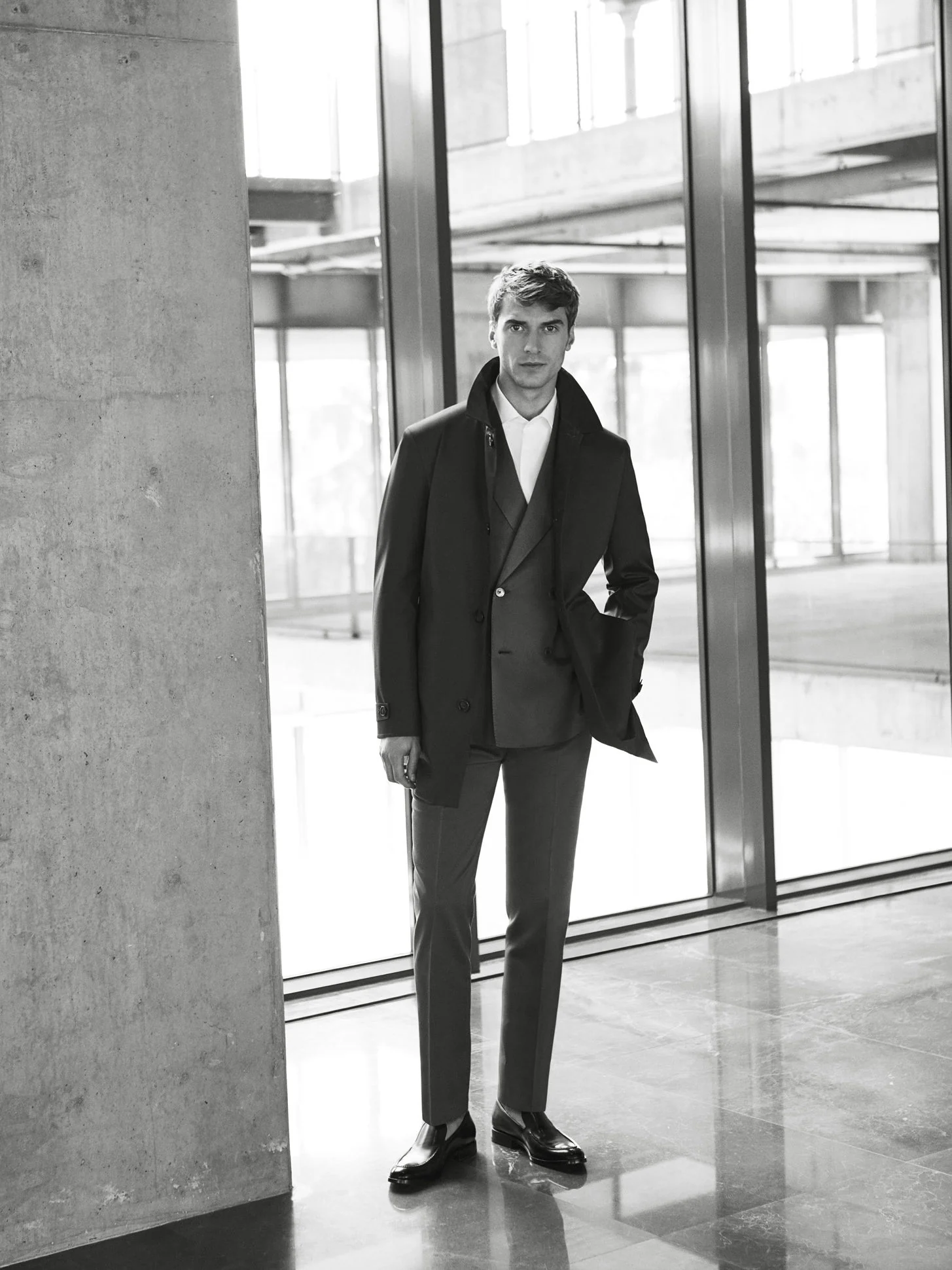 Black-and-white shot of a man in a dark overcoat and suit standing by a concrete pillar and glass wall, for Beymen.