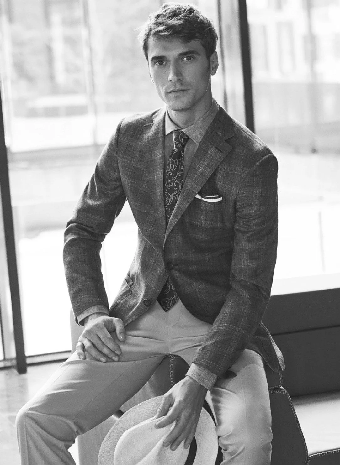 Black-and-white portrait of a man in a plaid blazer, paisley tie and light trousers holding a Panama hat, for Beymen.