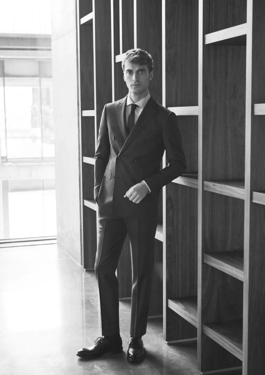 Black-and-white image of a man in a double-breasted suit and tie leaning on wooden shelving, for Beymen.