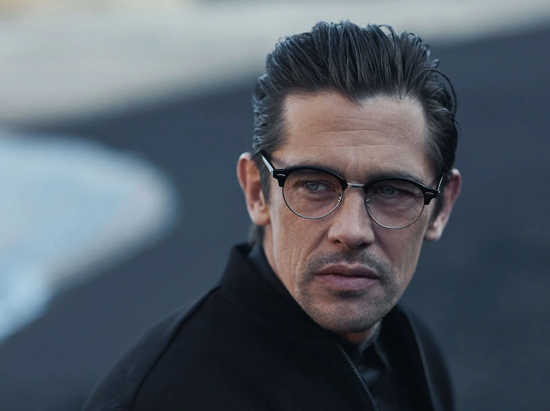 Man with slicked-back hair in browline glasses and black jacket on a dark beach, for Hiltl.