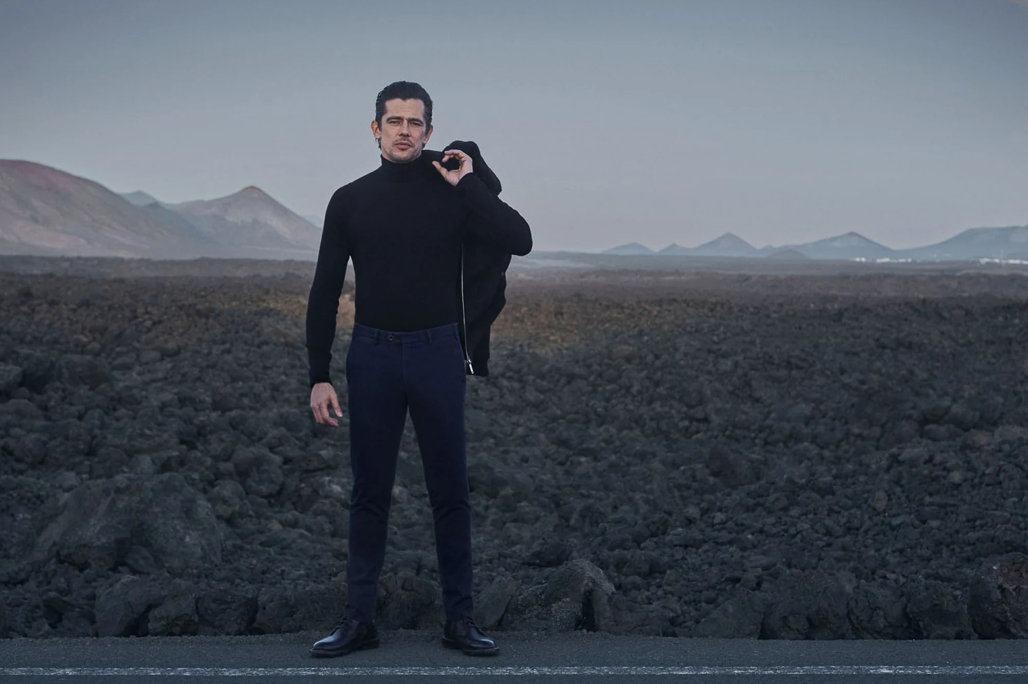 Man in black turtleneck and navy trousers holding a jacket over his shoulder on a volcanic lava field road, for Hiltl.