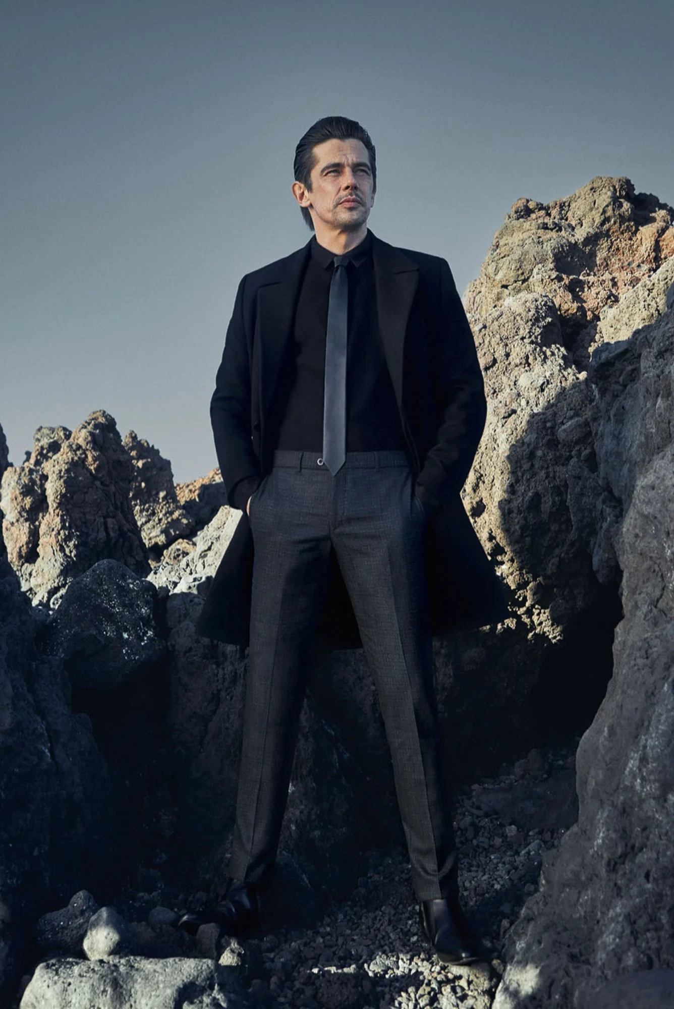 Man in black overcoat, black shirt and tie, and grey trousers standing among volcanic rocks under a clear sky, for Hiltl.