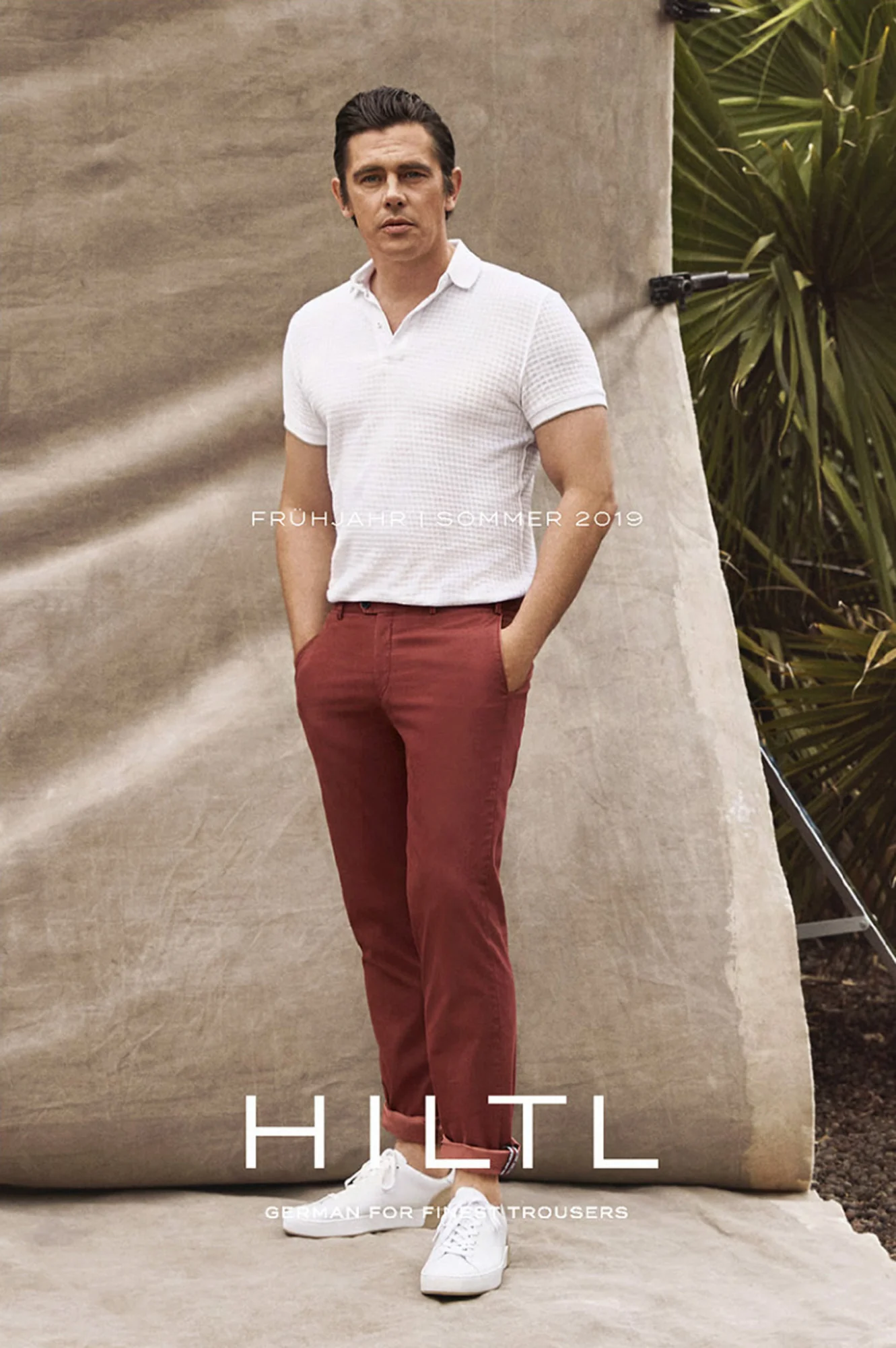 Man in a white knit polo and rust trousers standing before a canvas backdrop beside palms, for HILTL.