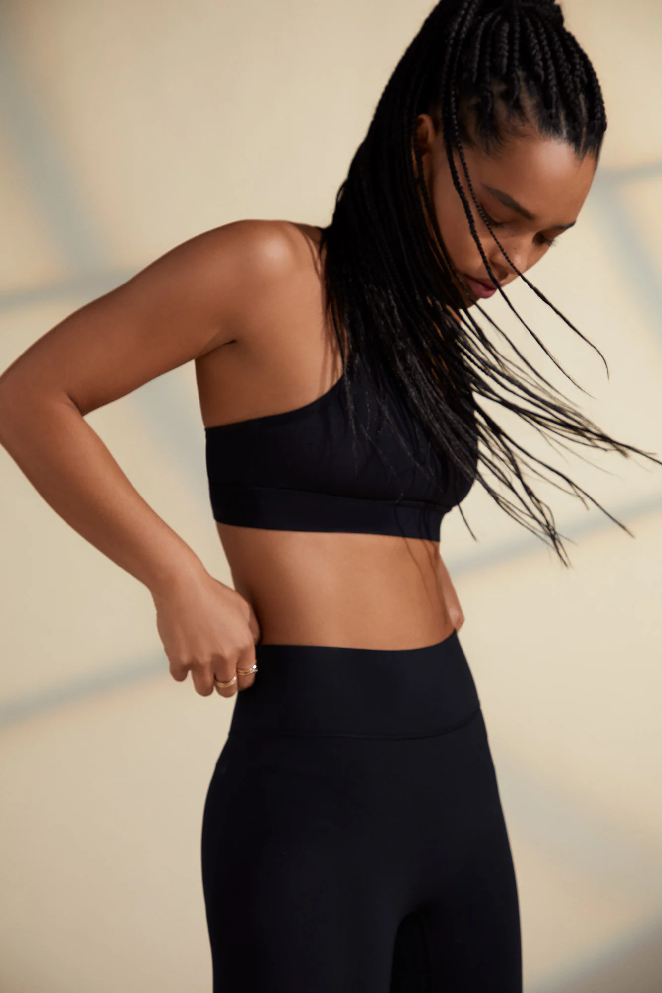 Model with long braids in a black sports bra and high-waist leggings against a warm beige wall, for Bandier.
