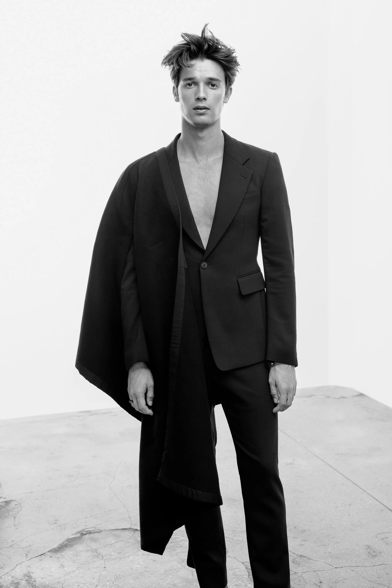 Black-and-white editorial of a male model in a black suit with a draped panel, bare chest, in a concrete studio, for Man About Town.