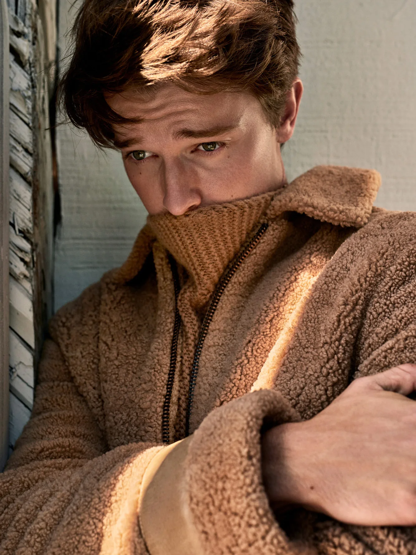 Young man in a camel teddy fleece coat and knit turtleneck pulled over his chin, sunlit editorial, for Man About Town.