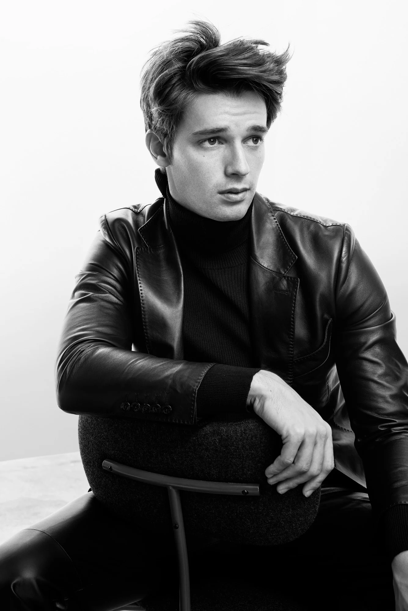 Black-and-white editorial portrait of a man in a leather blazer and turtleneck leaning on a chair, for Man About Town.