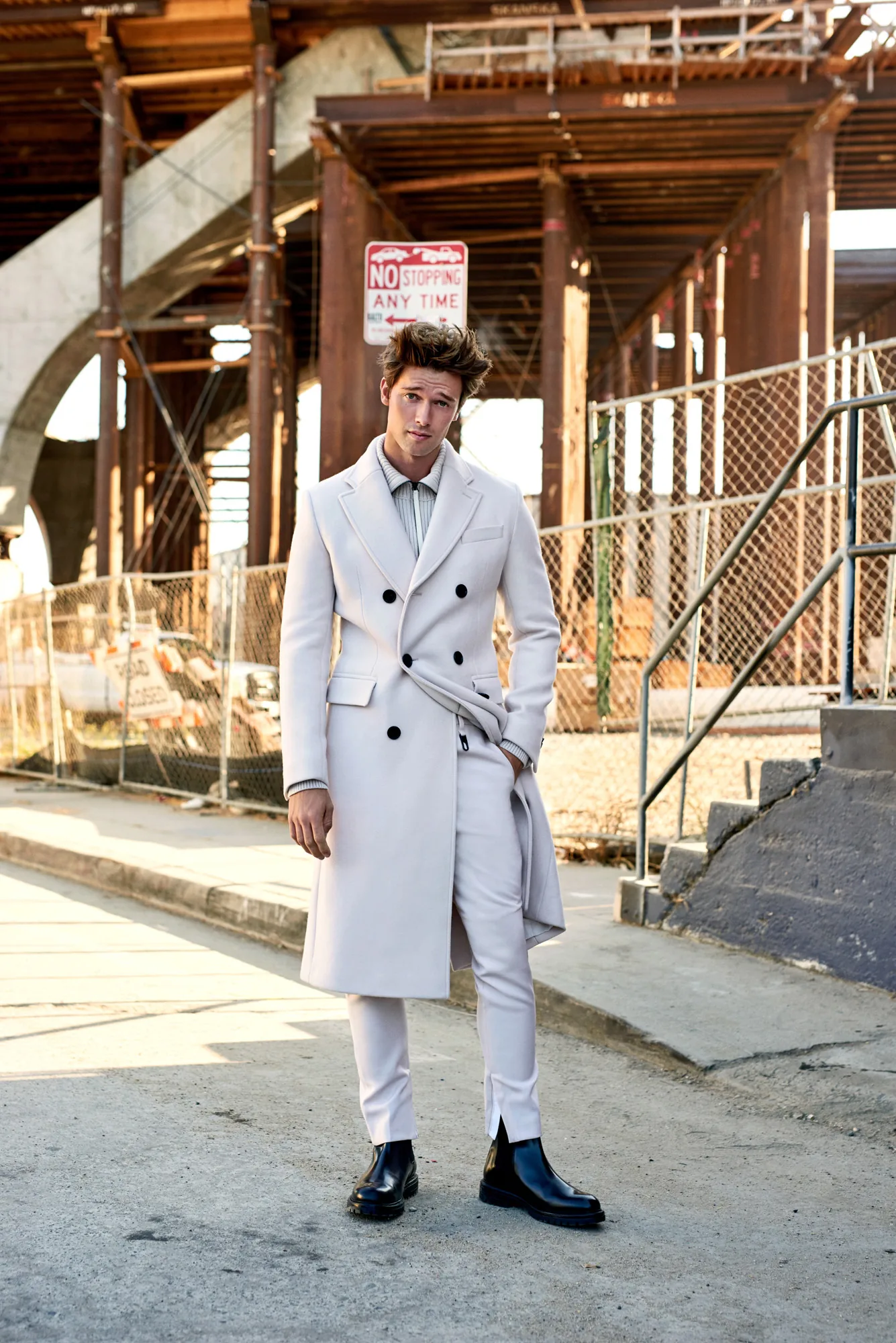 Man in a cream double-breasted overcoat and black boots stands under an elevated rail structure, for Man About Town.