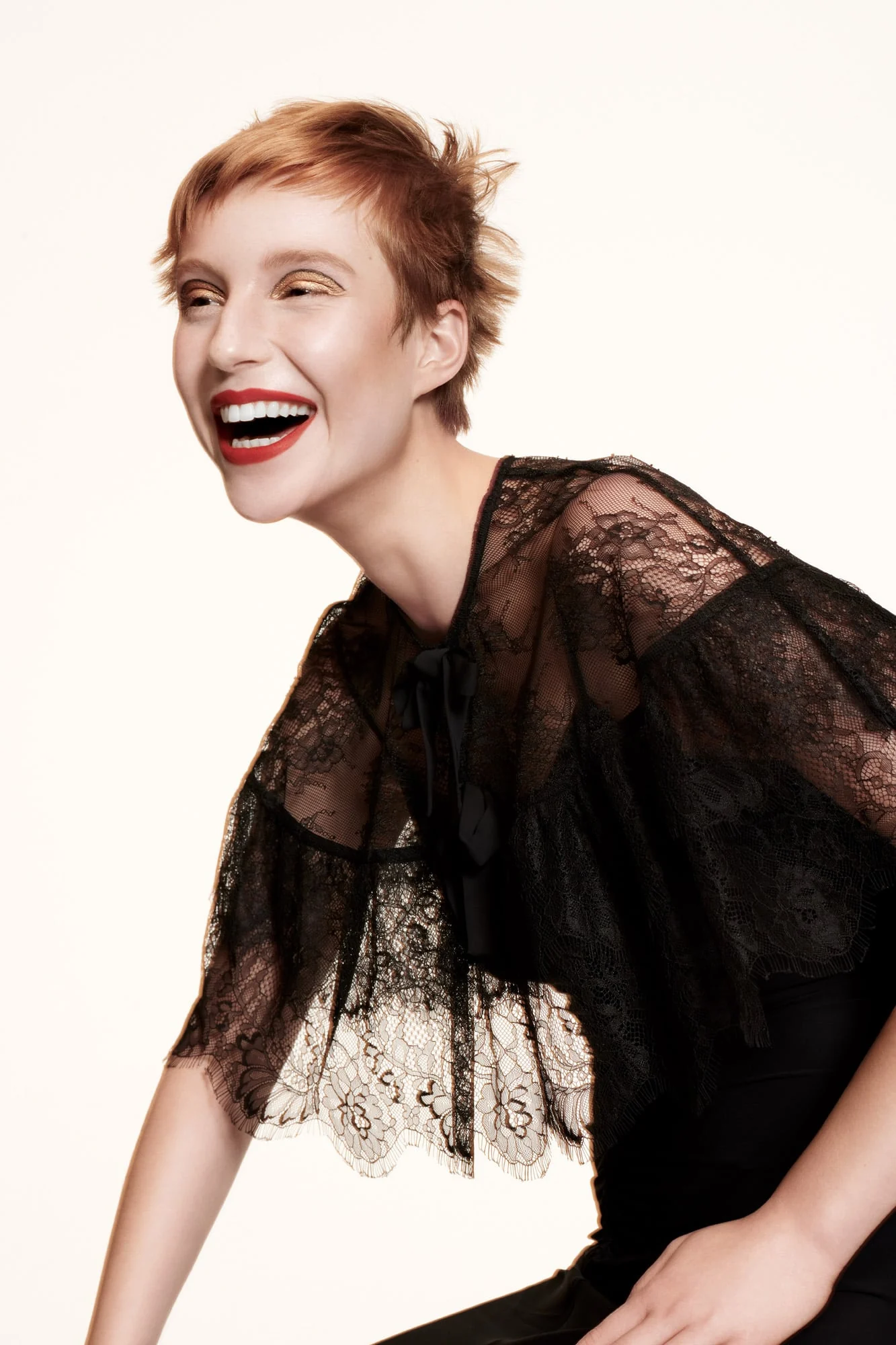 Laughing model with cropped copper hair and red lips in a sheer black lace cape on white, for Stella Magazine.