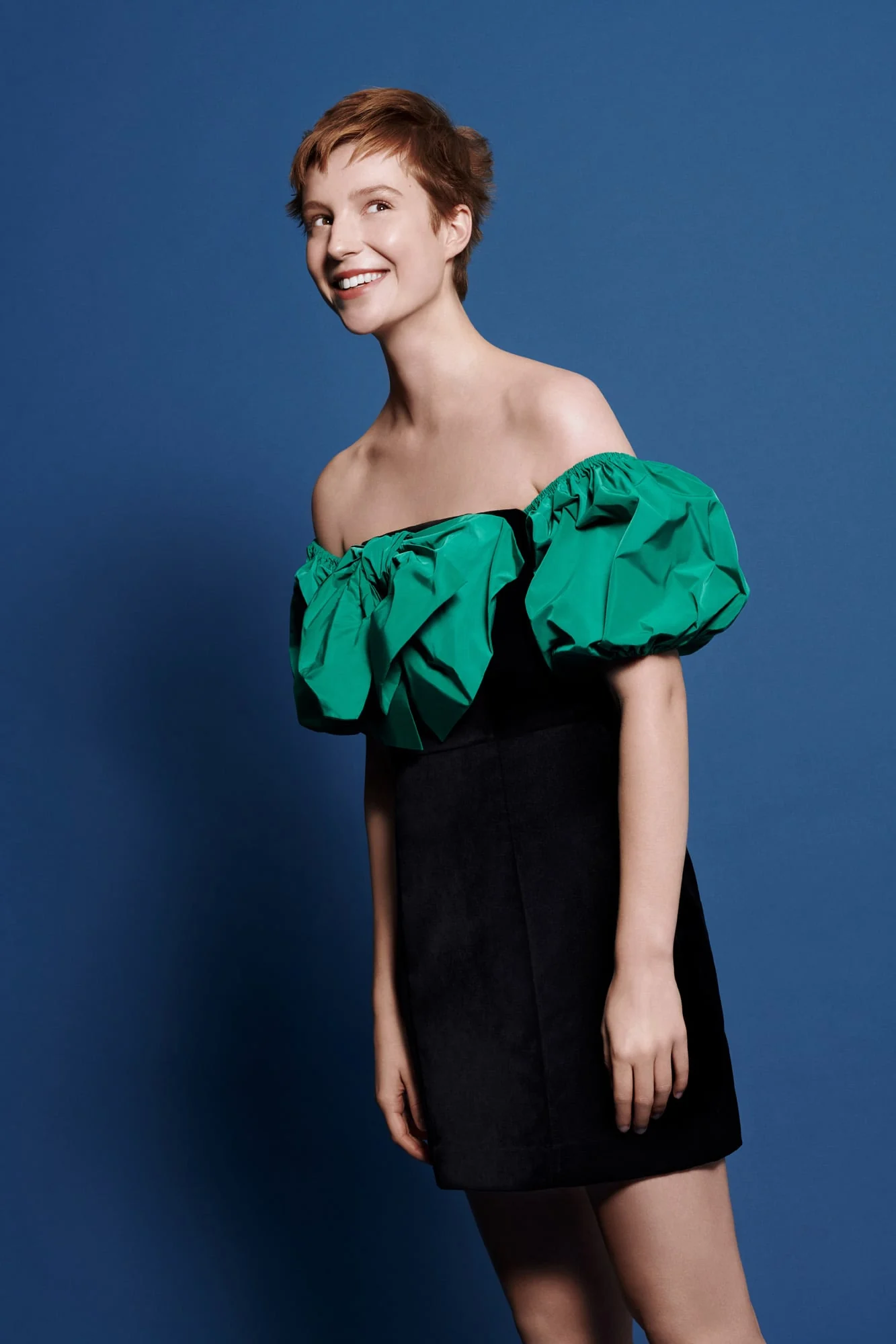 Smiling model with short red hair in a black mini dress with green off-shoulder puff sleeves on blue, for Stella Magazine.