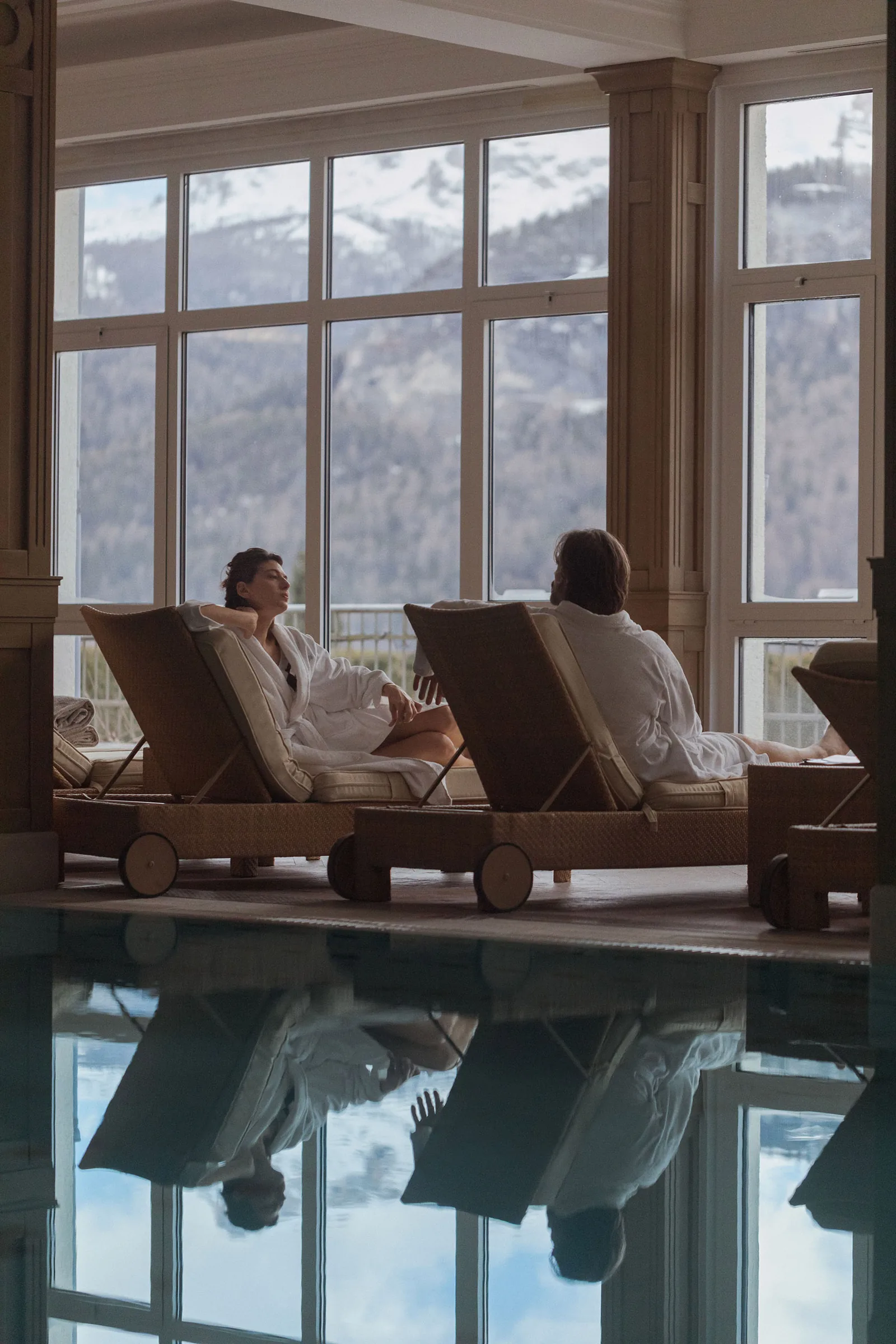 Two guests in white robes relax on loungers beside an indoor pool with mountain views, for Marriott.