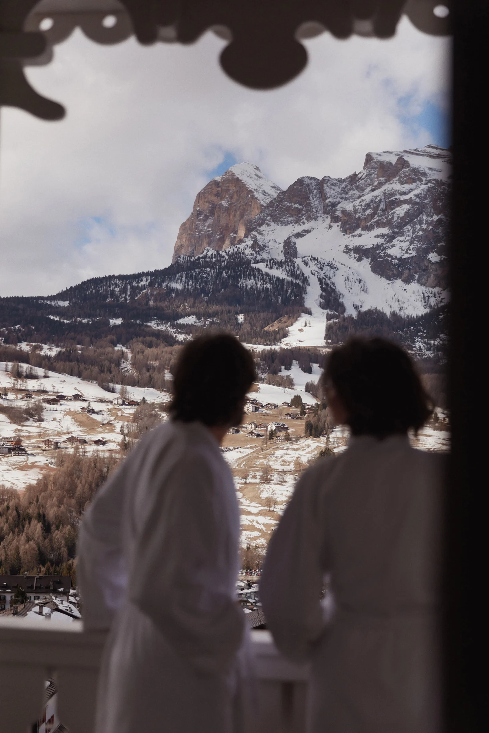 Two guests in white bathrobes on a balcony overlooking snowy Dolomite peaks and a village, for Marriott.