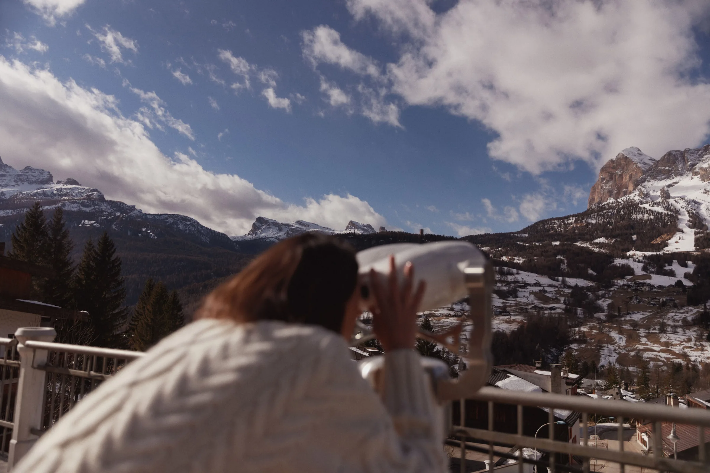 Woman in a cream knit sweater looks through a terrace viewer at snowy Dolomite peaks, for Marriott.