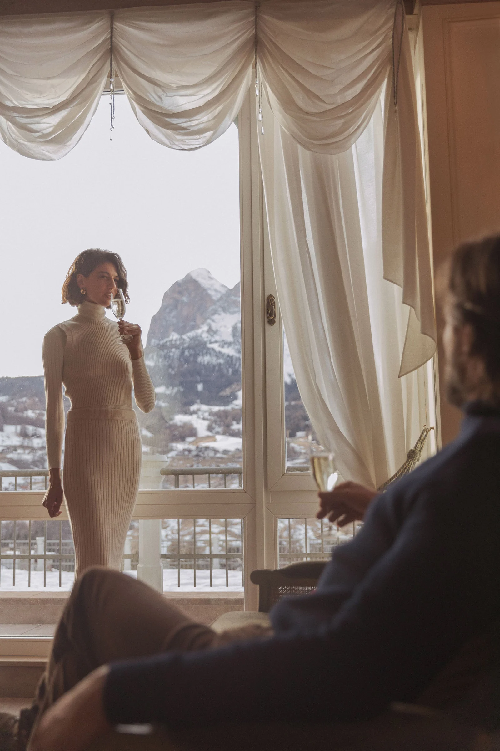 Woman in a knit dress sipping champagne by a hotel window with snowy mountains as a man watches, for Marriott.