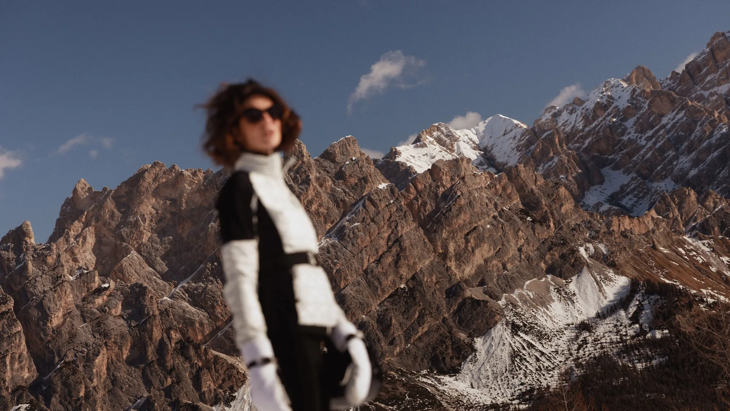 Woman in sunglasses and black-and-white ski suit, softly blurred before sharp snow-dusted rocky peaks, for Marriott.