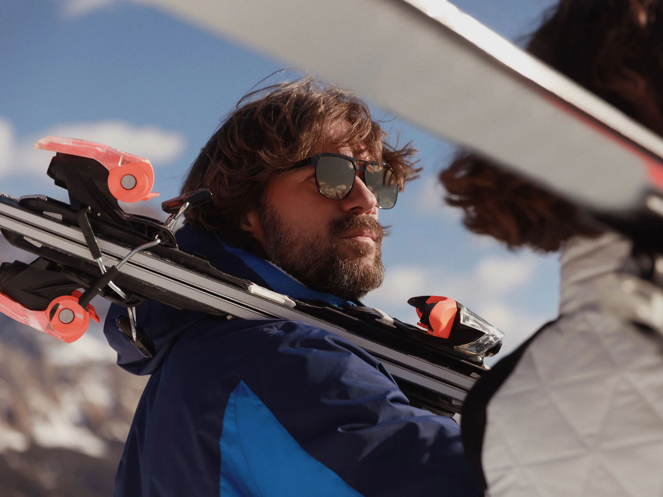 Bearded man in sunglasses and blue ski jacket carrying skis on his shoulder against a mountain sky, for Marriott.