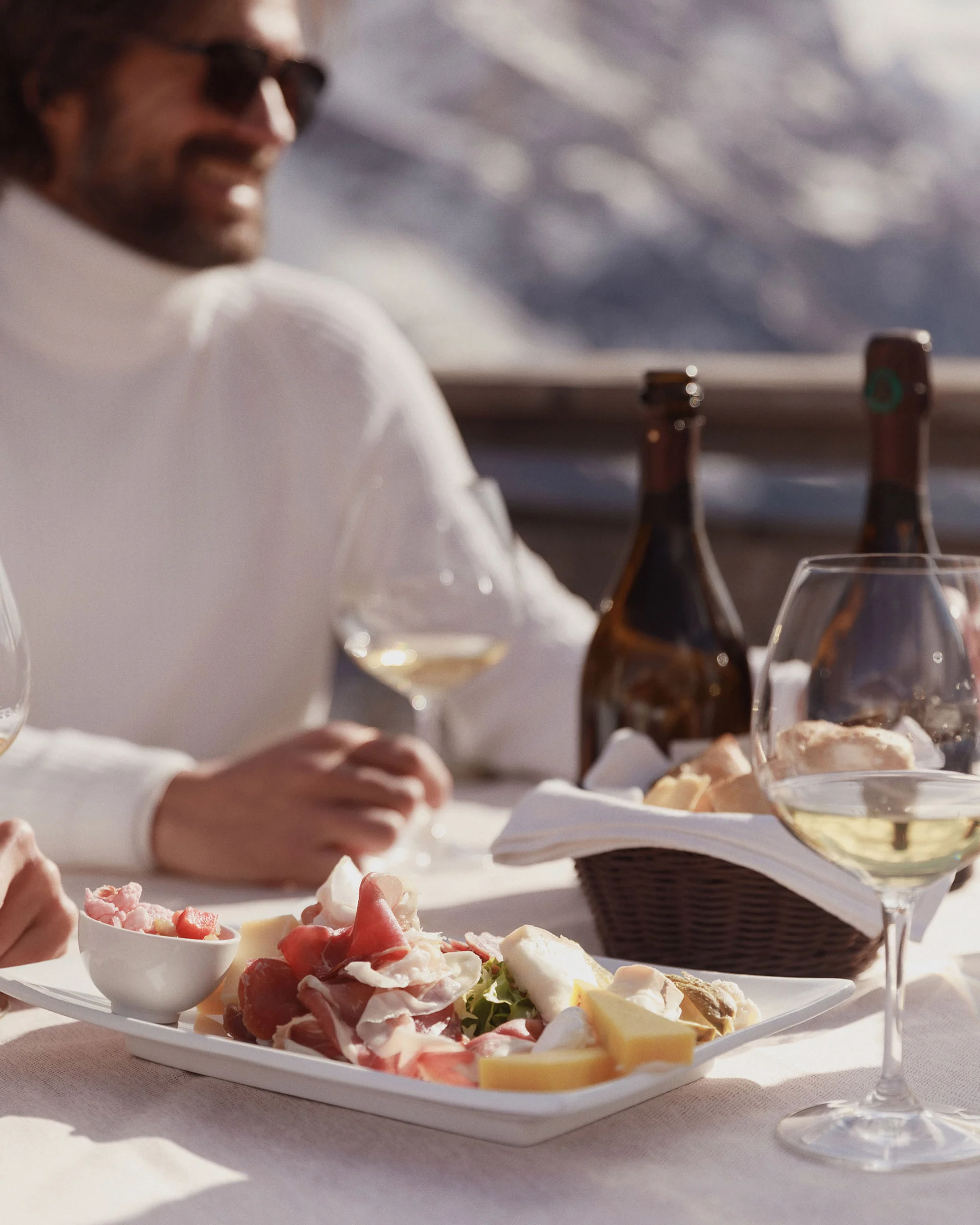 Man in white sweater at a sunny mountain terrace table with charcuterie, cheese, bread and white wine, for Marriott.