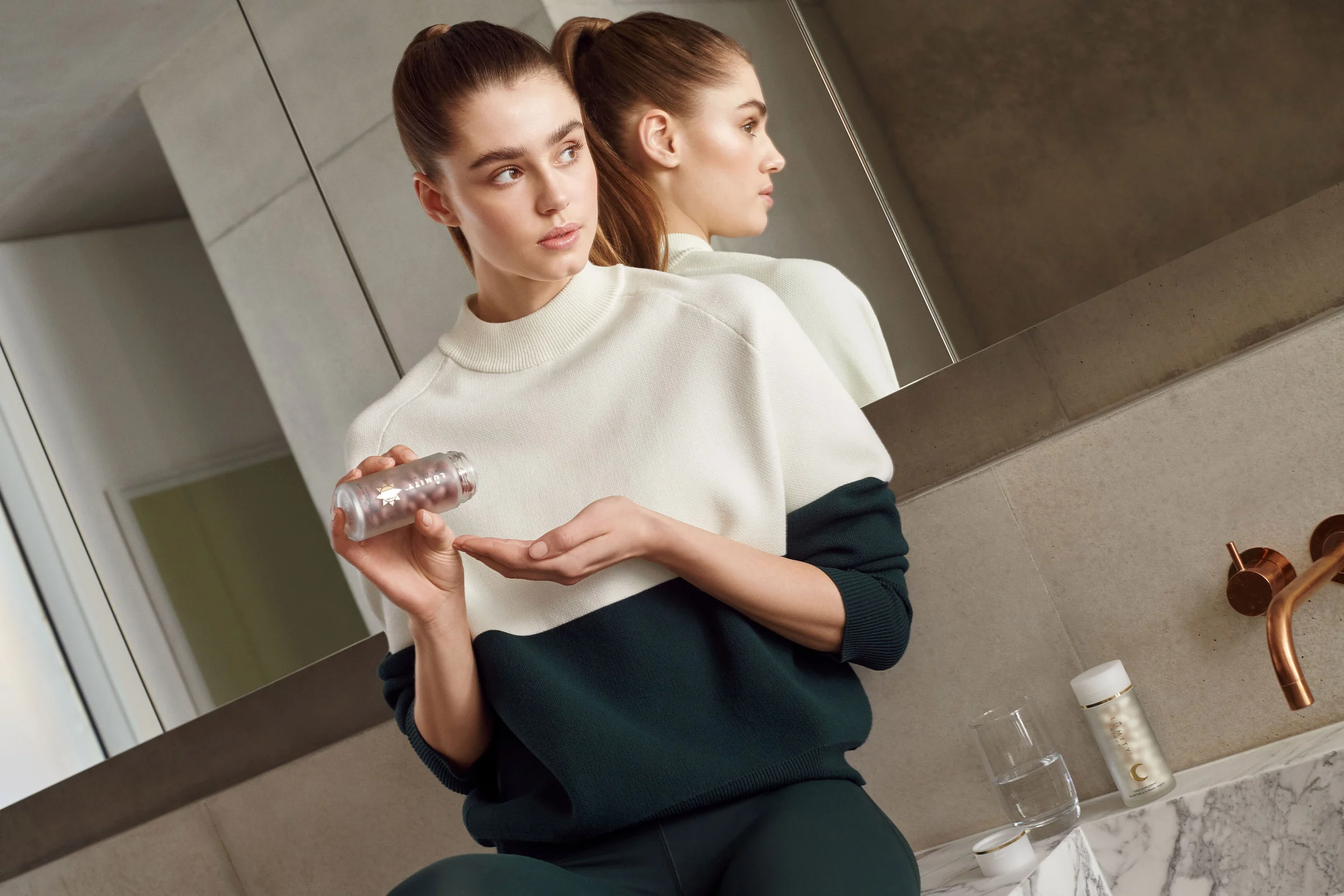 Model in a cream and green sweater holding a supplement bottle in a marble bathroom with copper taps, for Net-a-Porter.