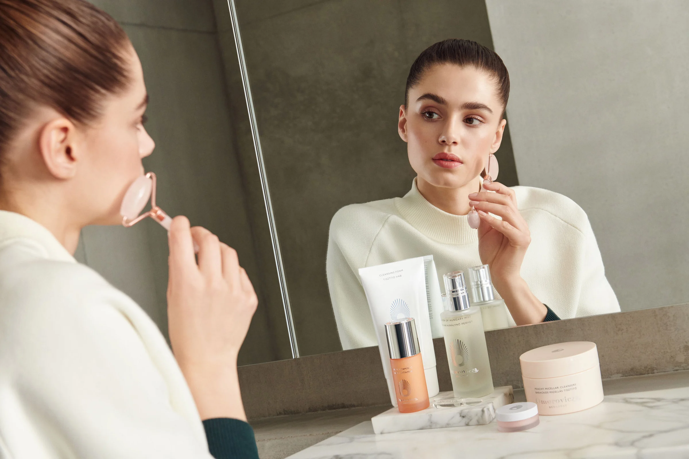 Woman using a rose quartz facial roller at a mirror beside skincare bottles on a marble counter, for Net-a-Porter.