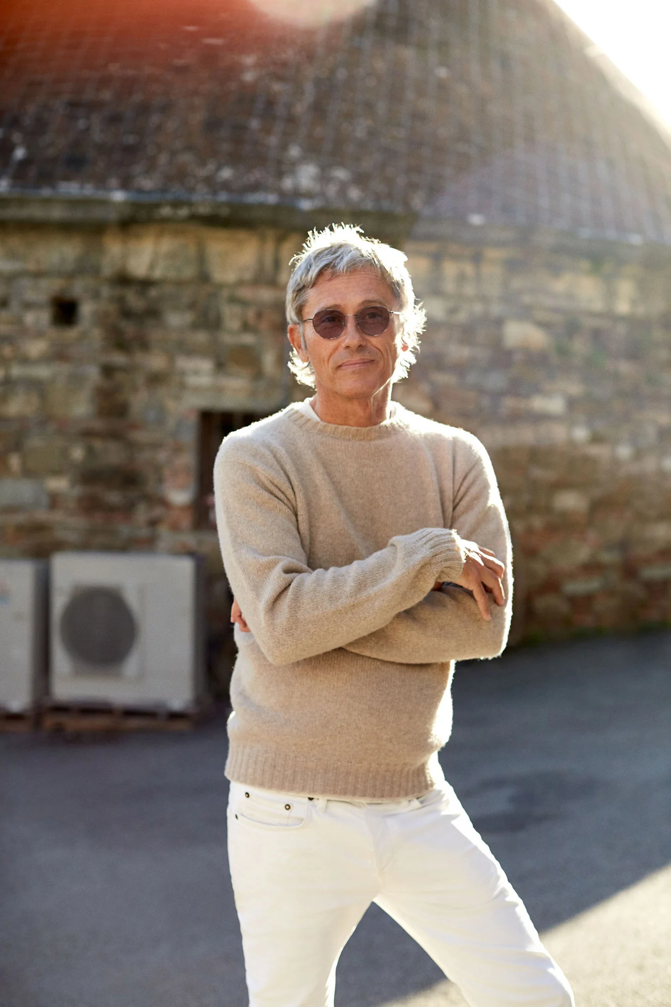 Grey-haired man in sunglasses, camel sweater and white jeans, arms crossed before a stone building, for The Sartorialist.