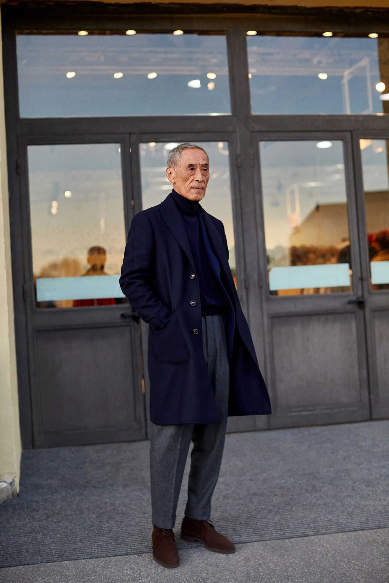Older man in a navy overcoat, turtleneck and grey trousers standing before glass doors, for The Sartorialist.