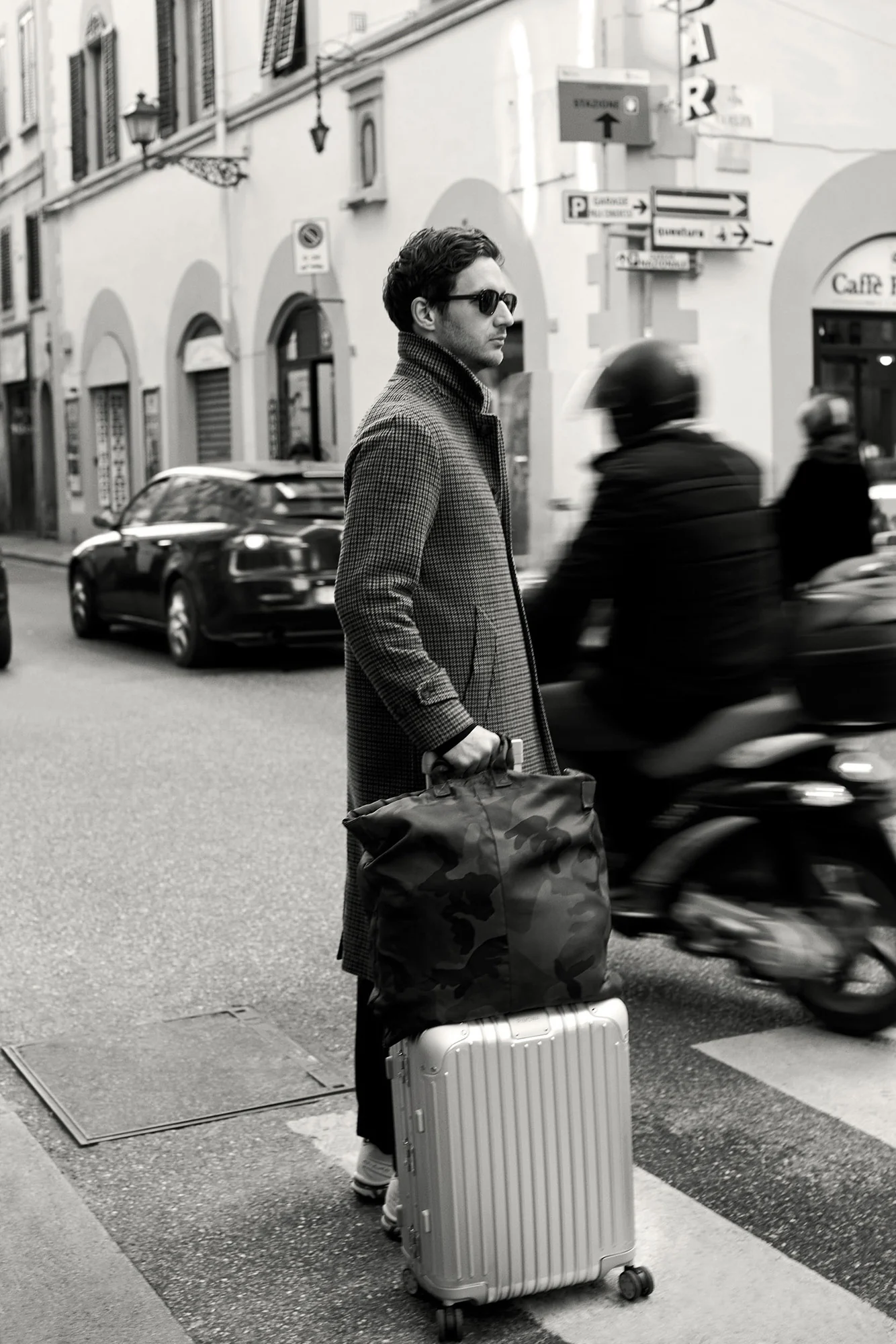 Black-and-white street photo of a man in a tweed coat with a camo bag and aluminum suitcase, for The Sartorialist.