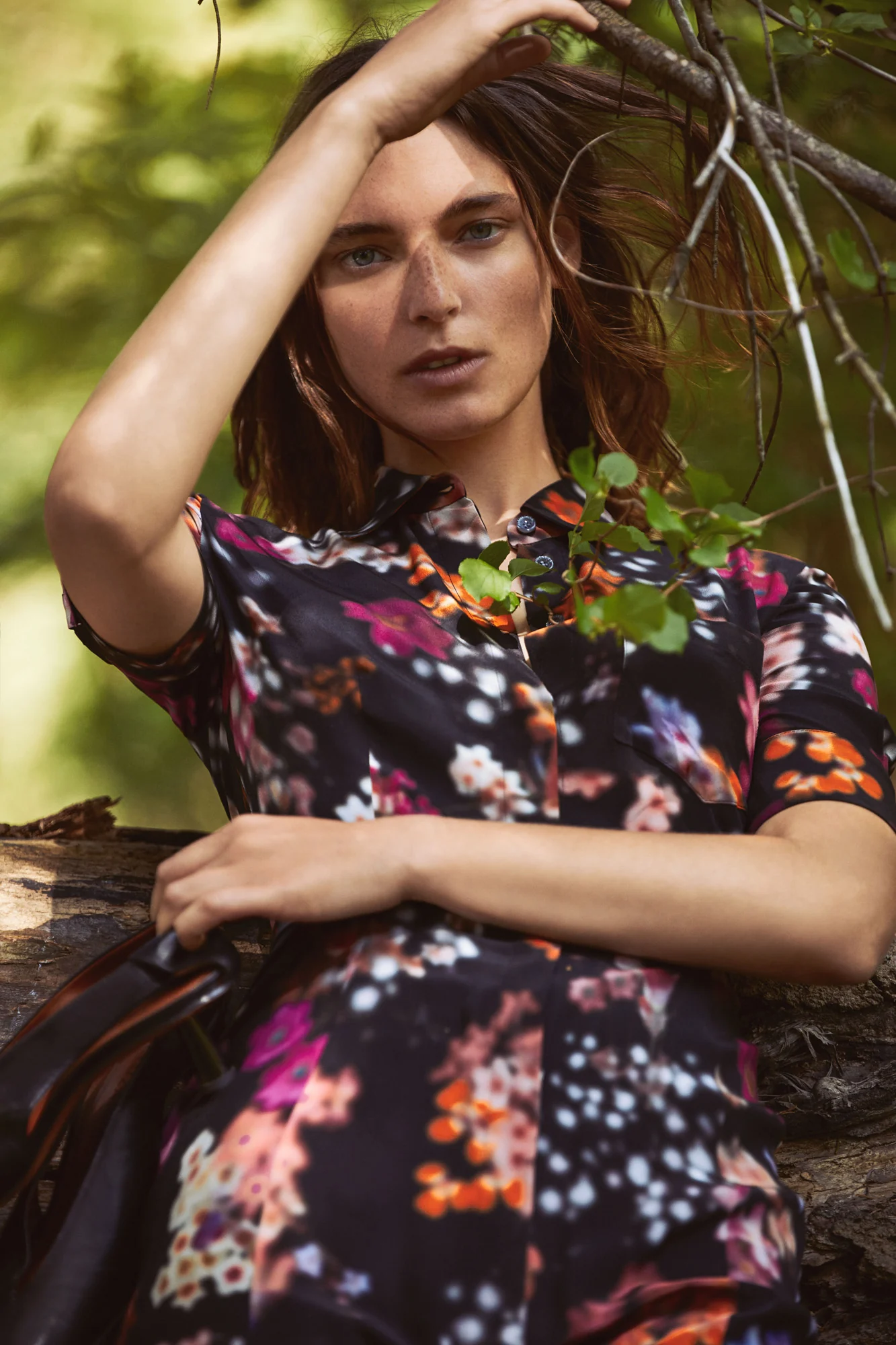 Model in a dark floral print shirt dress leaning on a log among green branches, hand in her hair, for Altuzarra.