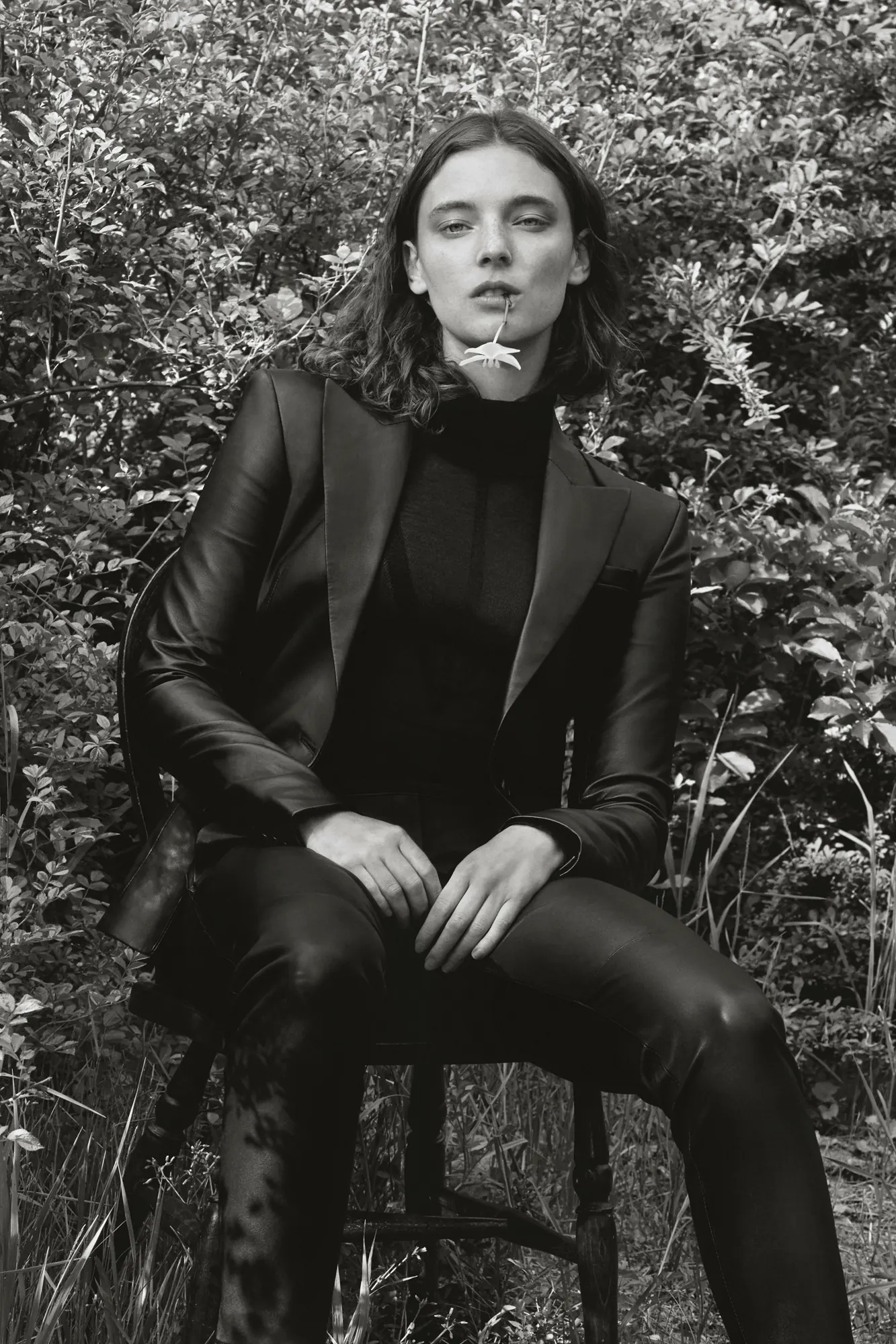 Black-and-white editorial of a model in a leather suit seated on a chair in a garden, a flower in her mouth, for Altuzarra.