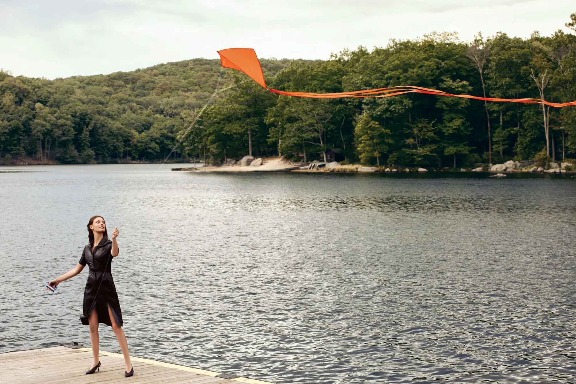Woman in a black dress flies an orange kite from a wooden dock on a forested lake, for Altuzarra.