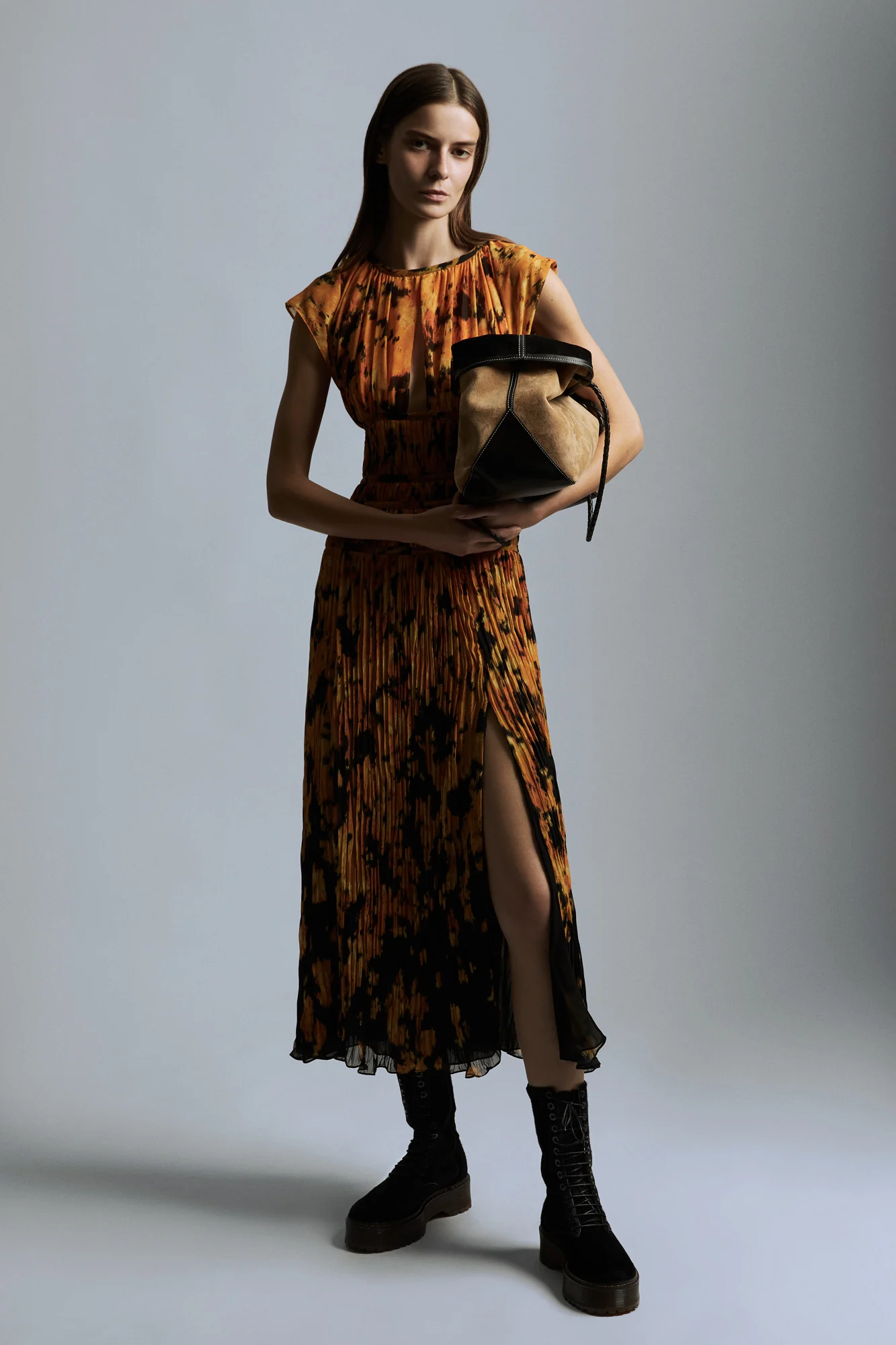 Model in an orange and black pleated midi dress with side slit, suede bag and lace-up boots on grey, for Altuzarra.