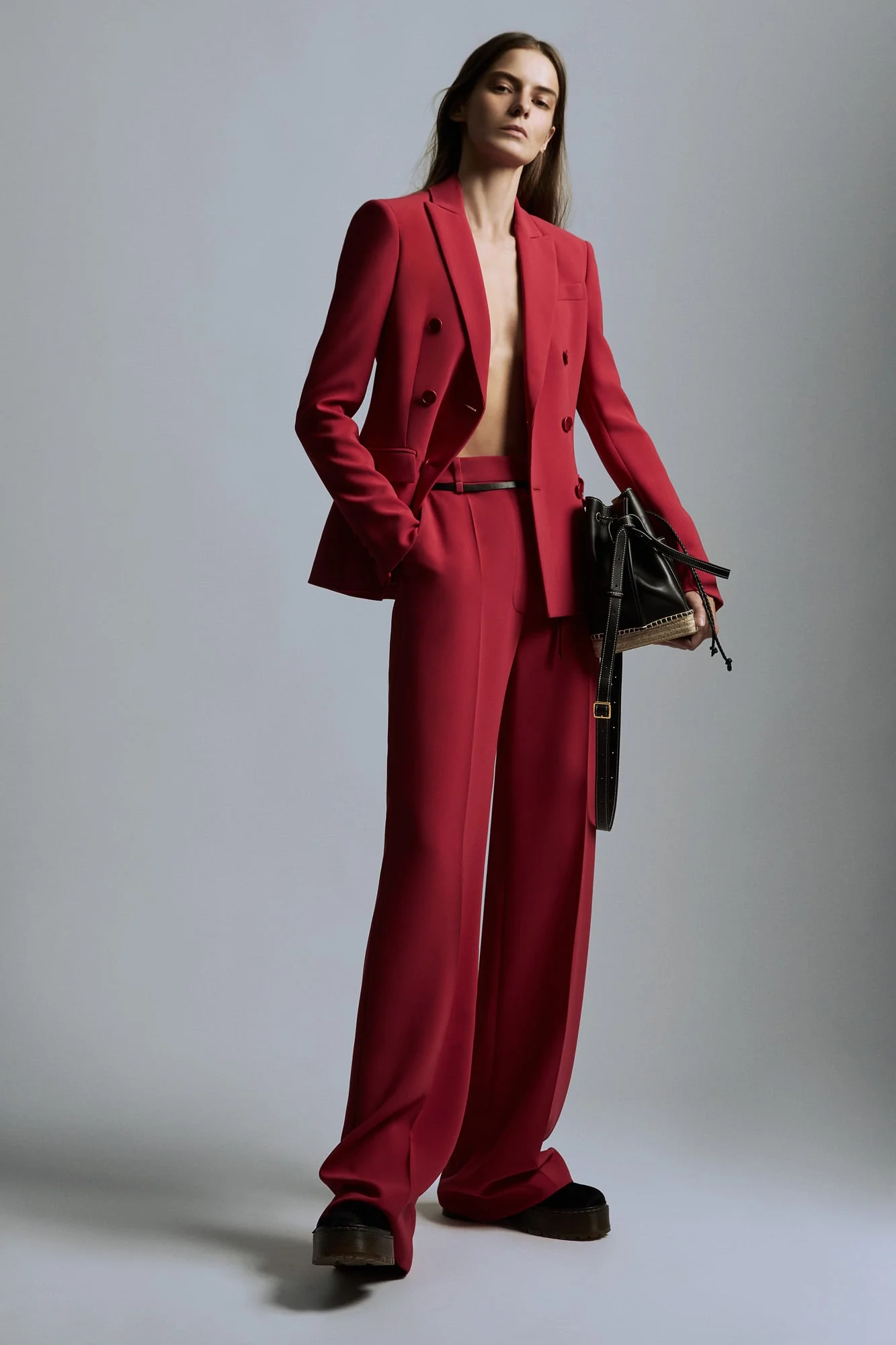 Model in a red double-breasted blazer and wide-leg trousers holding a black bucket bag on grey backdrop, for Altuzarra.