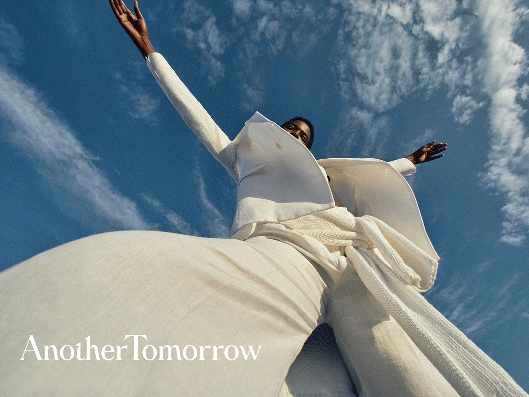Low-angle view of a model in a white linen suit and knit scarf, arms raised against a blue sky, for Another Tomorrow.