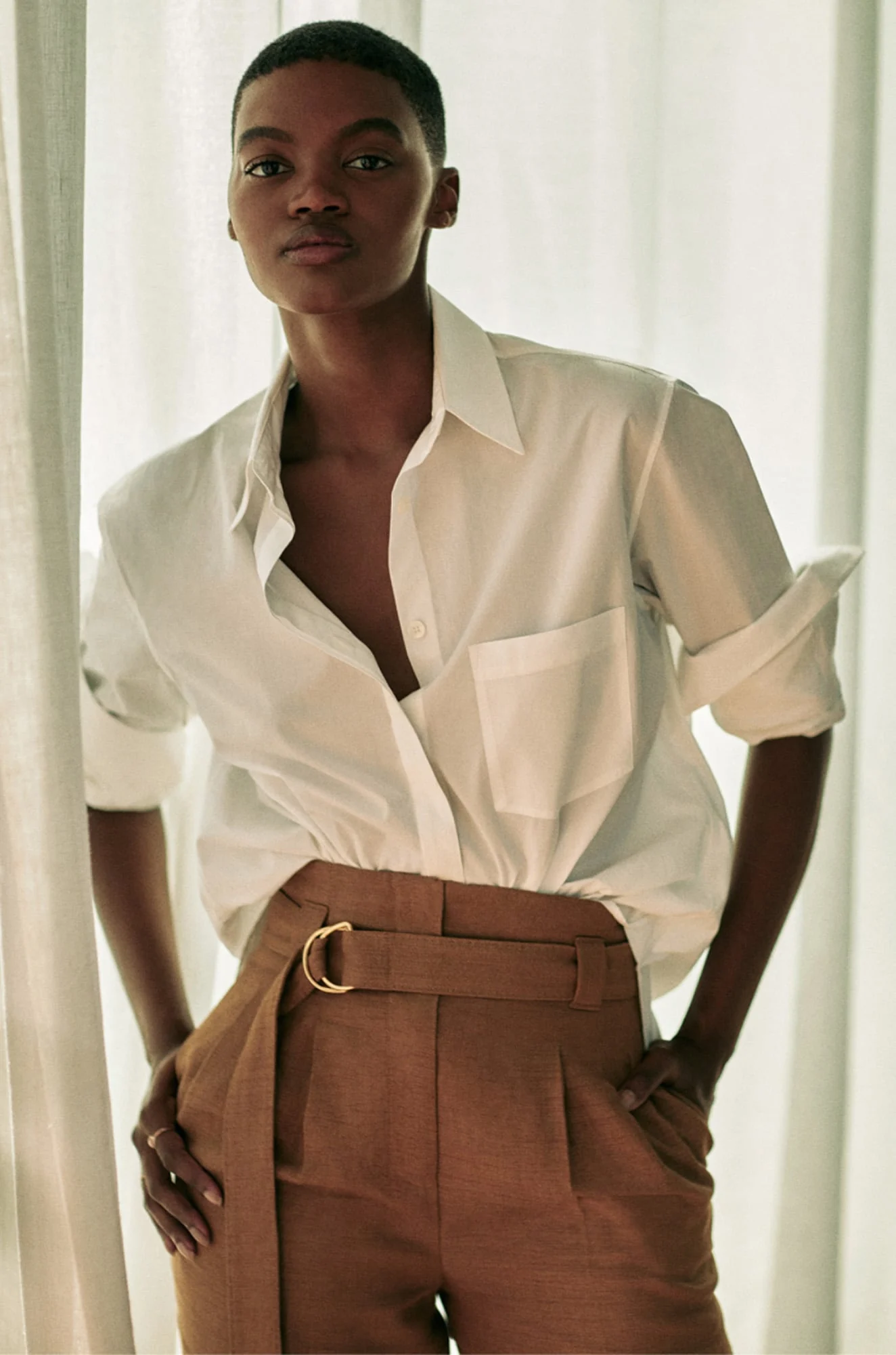 Model in an oversized white shirt and belted camel trousers posing by sheer curtains, for Another Tomorrow.