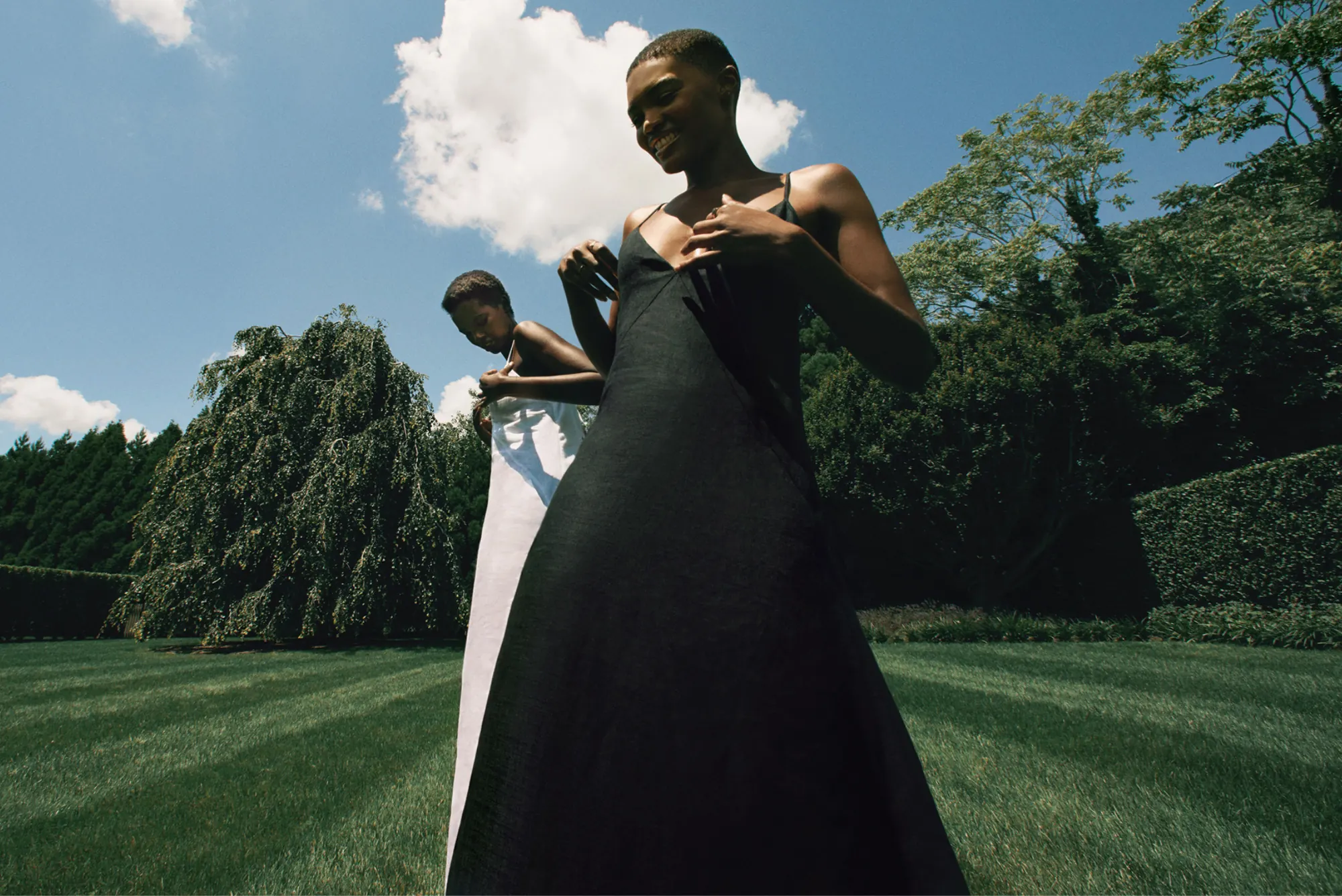 Two models in black and white slip dresses on a striped lawn under blue sky, for Another Tomorrow.