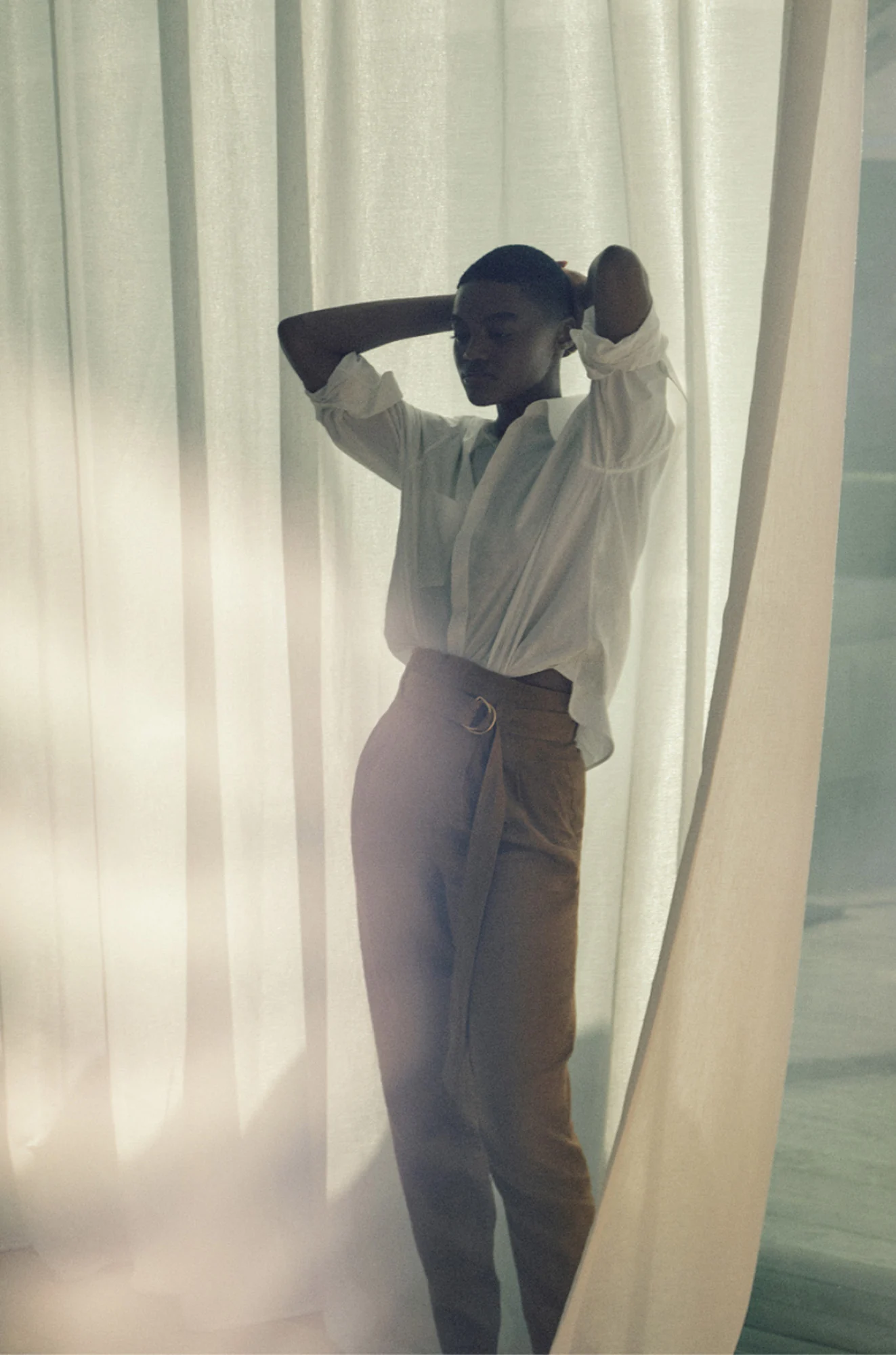 Model in a white shirt and belted tan trousers posing among sheer white curtains in soft light, for Another Tomorrow.