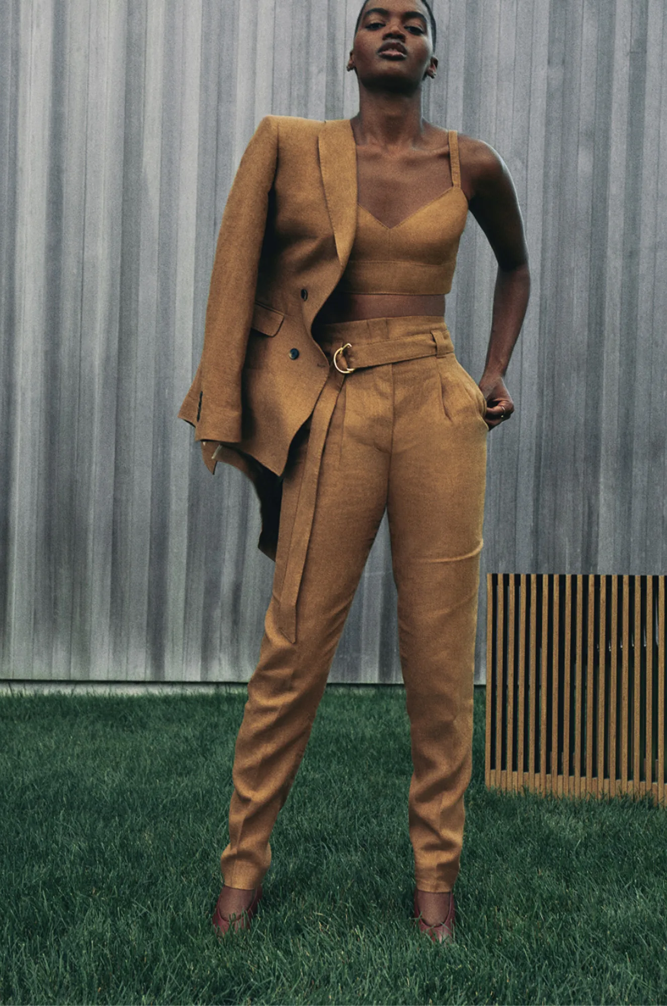 Model in a camel linen blazer, bralette and belted trousers on grass by a grey wall, for Another Tomorrow.
