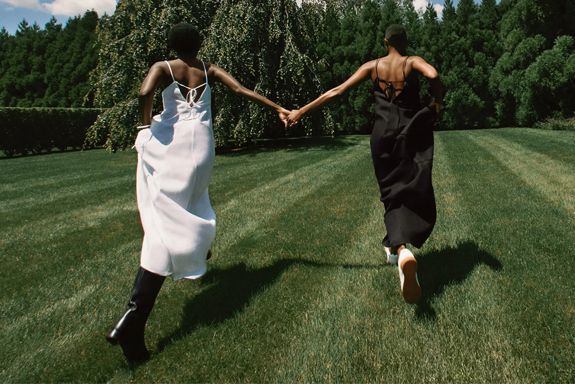 Two models in white and black slip dresses run hand in hand across a striped green lawn, for Another Tomorrow.