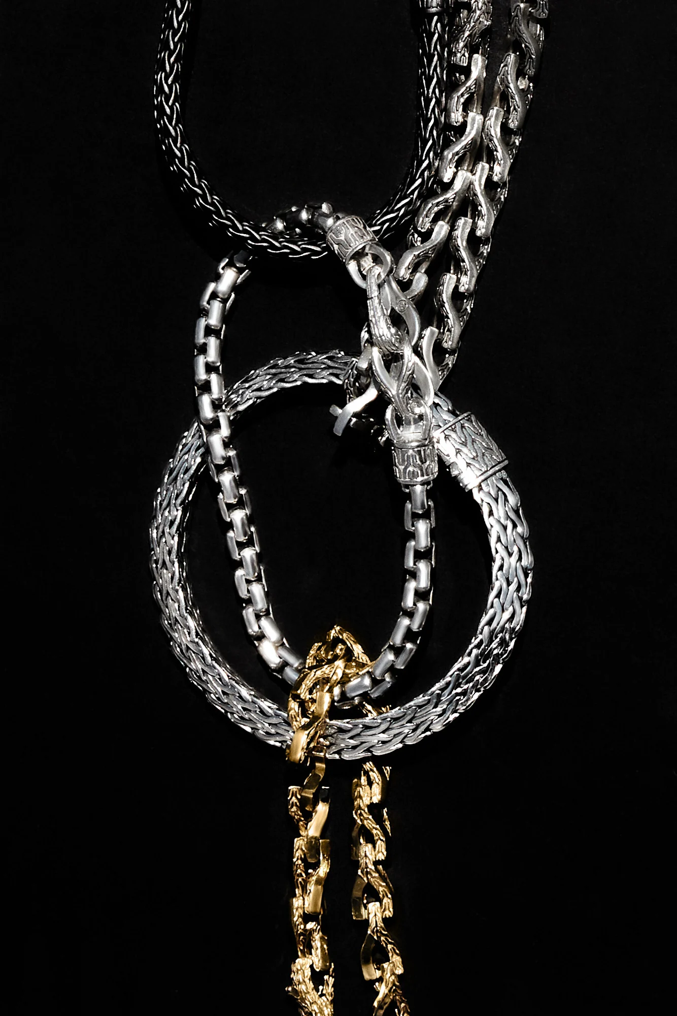 Silver and gold chain necklaces and bracelets looped together against a black background, for John Hardy.