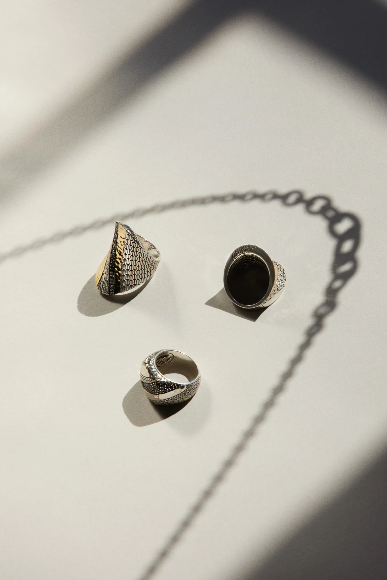 Three silver rings with gold, black stone and chain textures on a white surface beside a chain's shadow, for John Hardy.