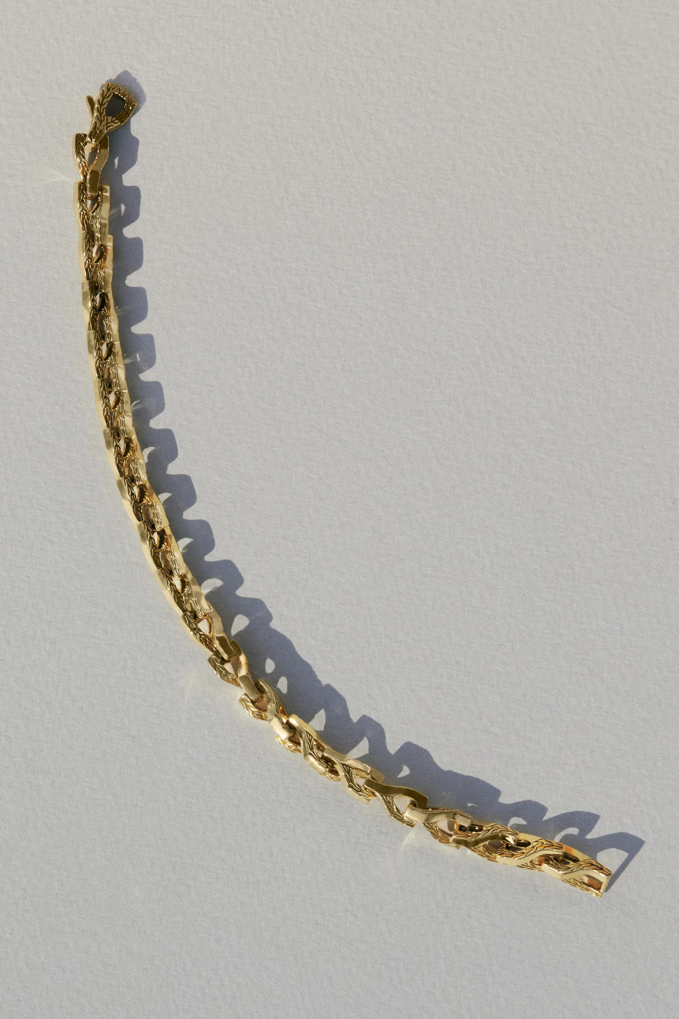 Gold link bracelet with carved texture lying on a white textured surface, casting a sharp shadow, for John Hardy.