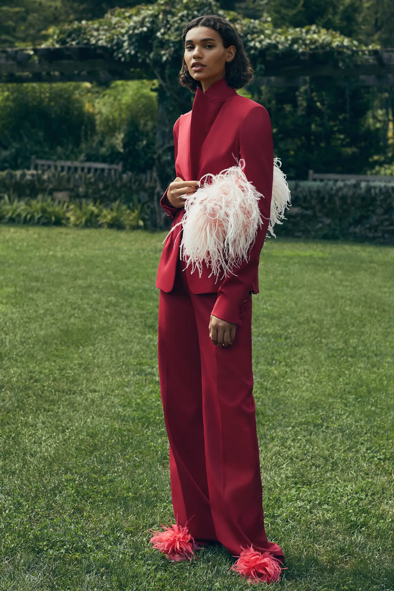 Model in a red trouser suit with pink ostrich feather cuffs and slippers on a garden lawn, Fall 2020 campaign, for Altuzarra.