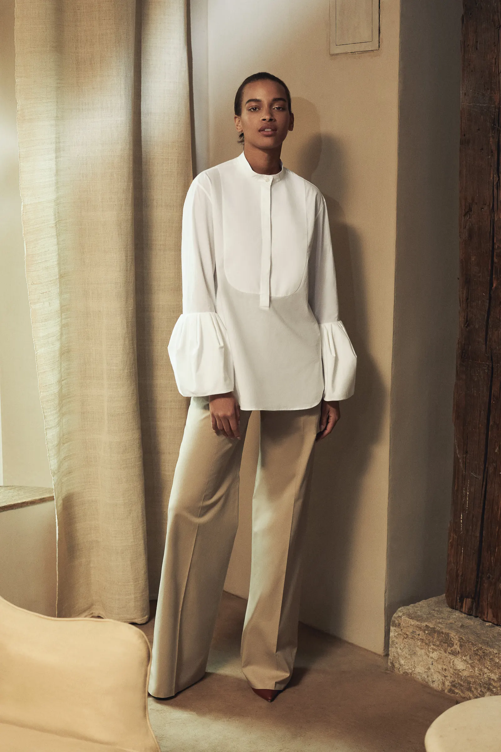 Model in a white bell-sleeve bib shirt and wide beige trousers beside linen curtains, for Another Tomorrow.
