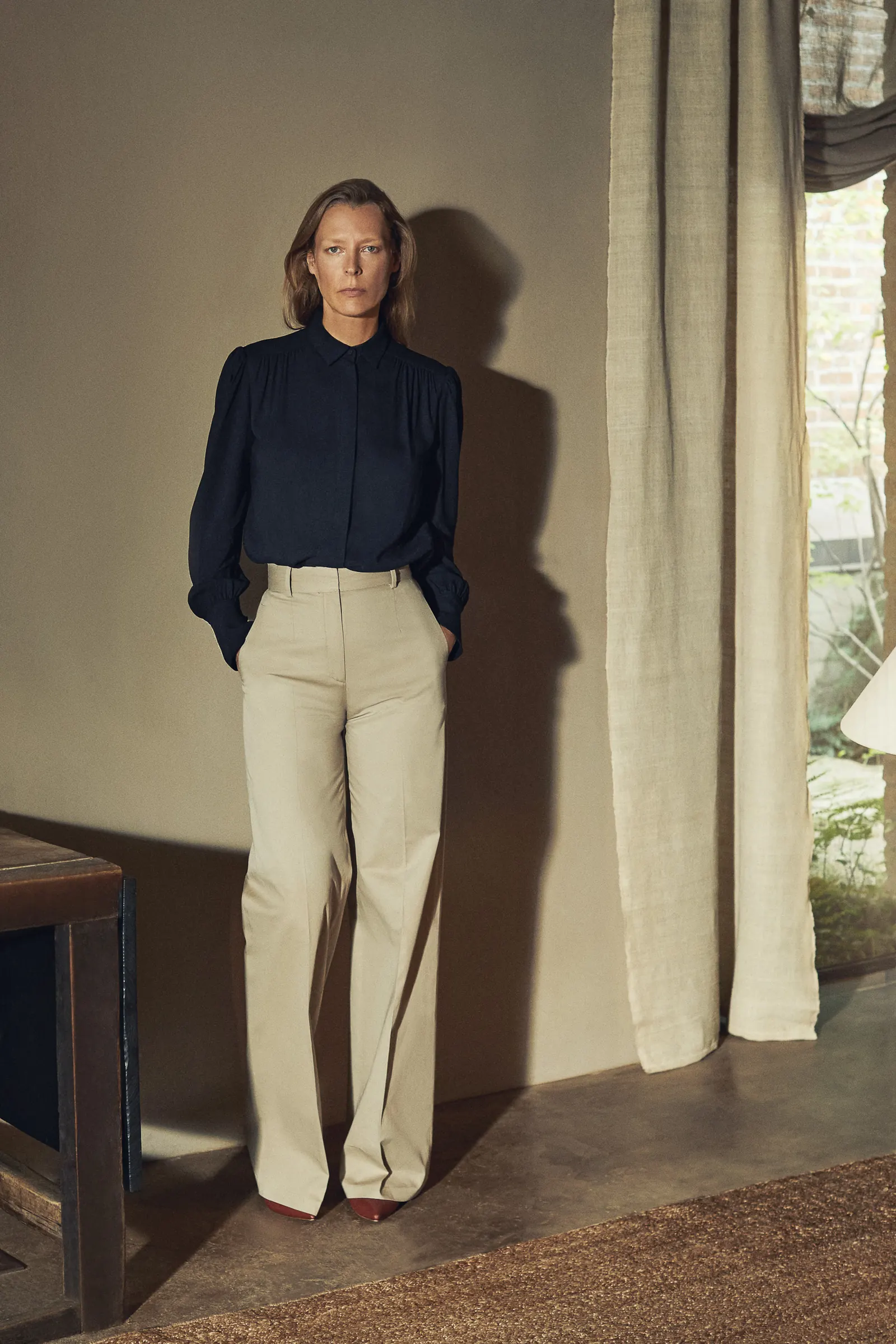 Woman in a navy blouse and wide-leg khaki trousers standing by a linen curtain in a warm interior, for Another Tomorrow.